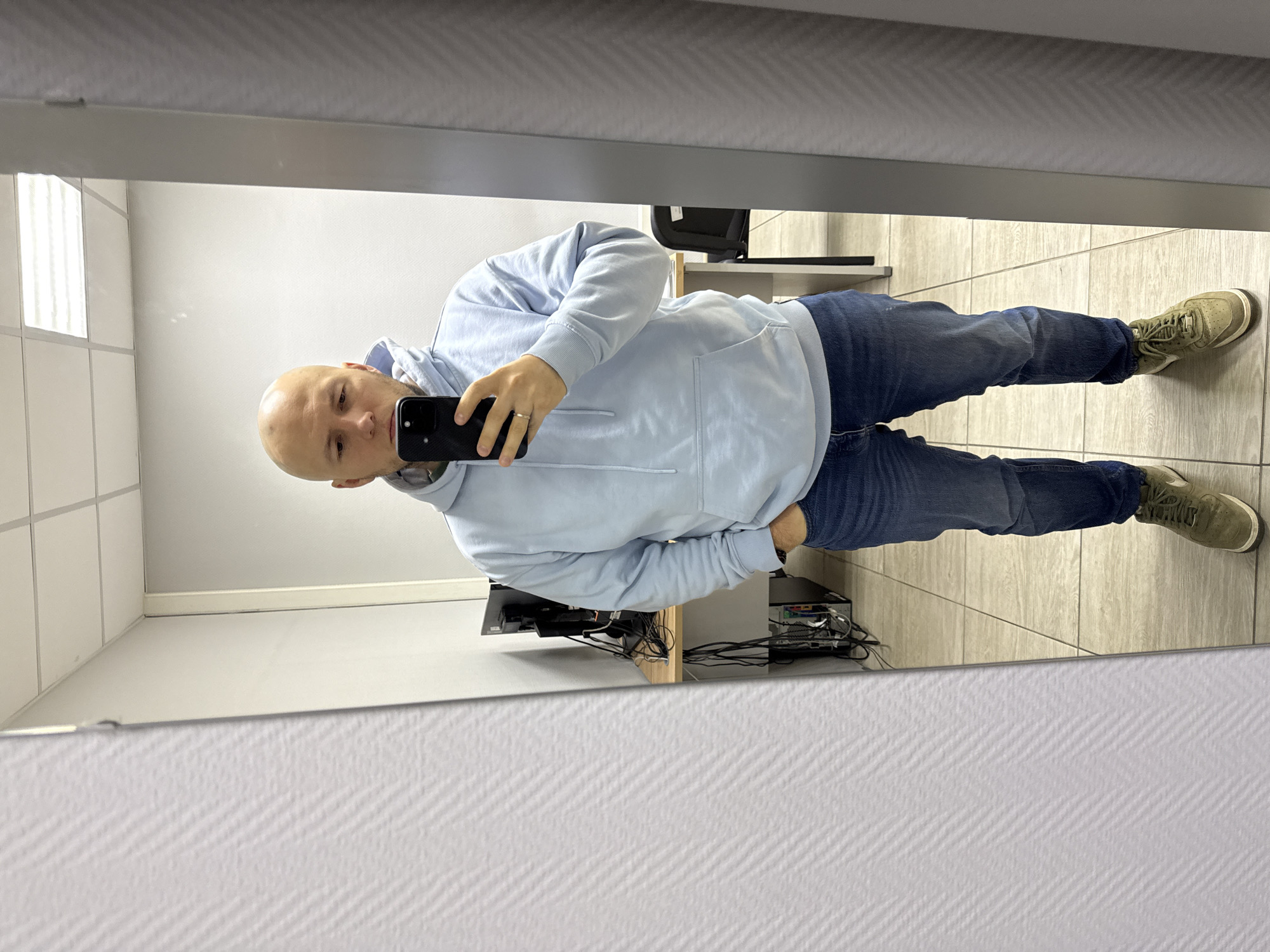 A bald man stands in front of a mirror, holding a smartphone to take a picture of himself. He wears a light blue hoodie, blue jeans, and green sneakers. The location appears to be an office space in Krasnoyarsk, Russia, with tiled floors, white walls, and a desk with computer equipment. A chair is visible in the background.