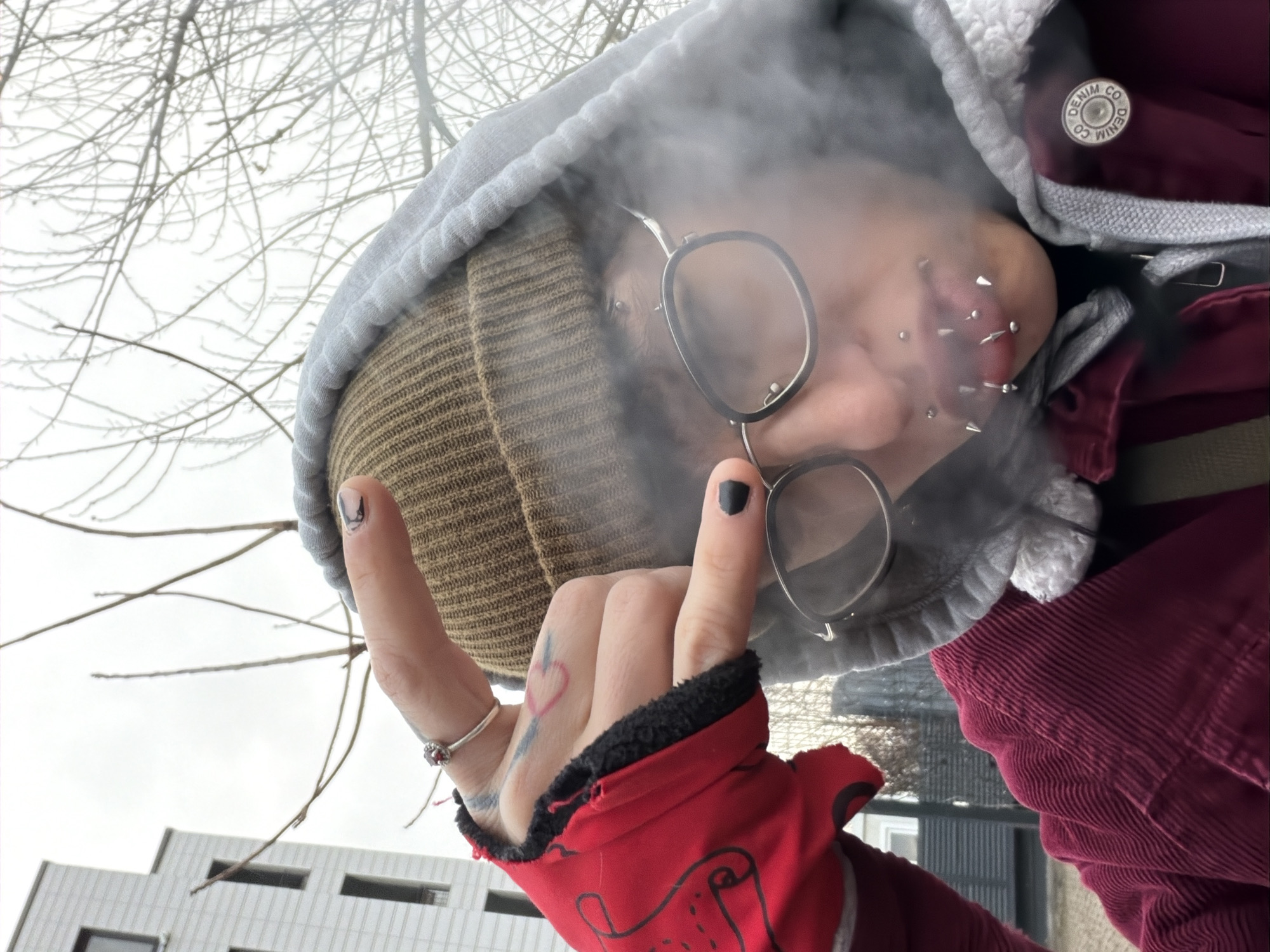 The image shows a person wearing a brown beanie, a gray hooded sweatshirt, glasses, and a red corduroy jacket. The person has numerous piercings around their mouth. They are holding up their hand with a gesture resembling devil horns. They have a black nail polish and a heart tattoo on their hand. Smoke or fog is surrounding their face. The background shows bare trees and a white building with windows. The photo was taken in the city of Sofia, Bulgaria.