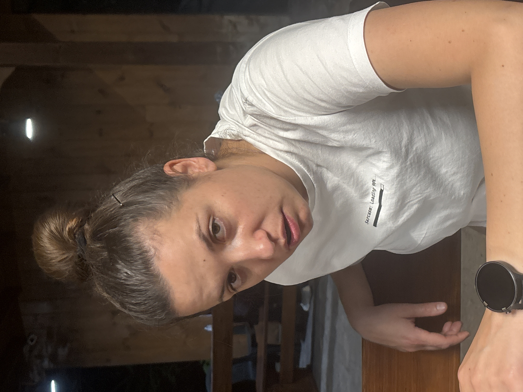 A woman with her hair pulled back in a bun is looking directly at the camera. She is wearing a white t-shirt. A smartwatch is visible on her left wrist. The background appears to be an interior space with wood paneling. The location in Sofia, Bulgaria cannot be determined from the image.