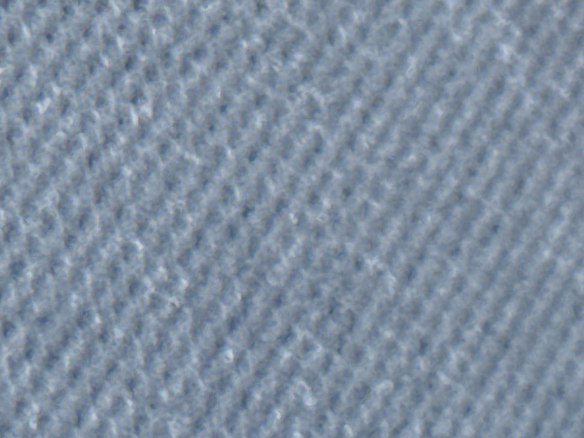 The image is a close-up, macro photograph of a light blue, textured fabric. The texture appears to be a knit or woven pattern with diagonal lines formed by the interlacing of threads. There are subtle variations in the shading, suggesting a slightly uneven surface or lighting. The image is abstract, focusing entirely on the material's surface. There is no discernible setting, people, or objects within the frame that would suggest a specific location in Varna, Bulgaria, or any other place.