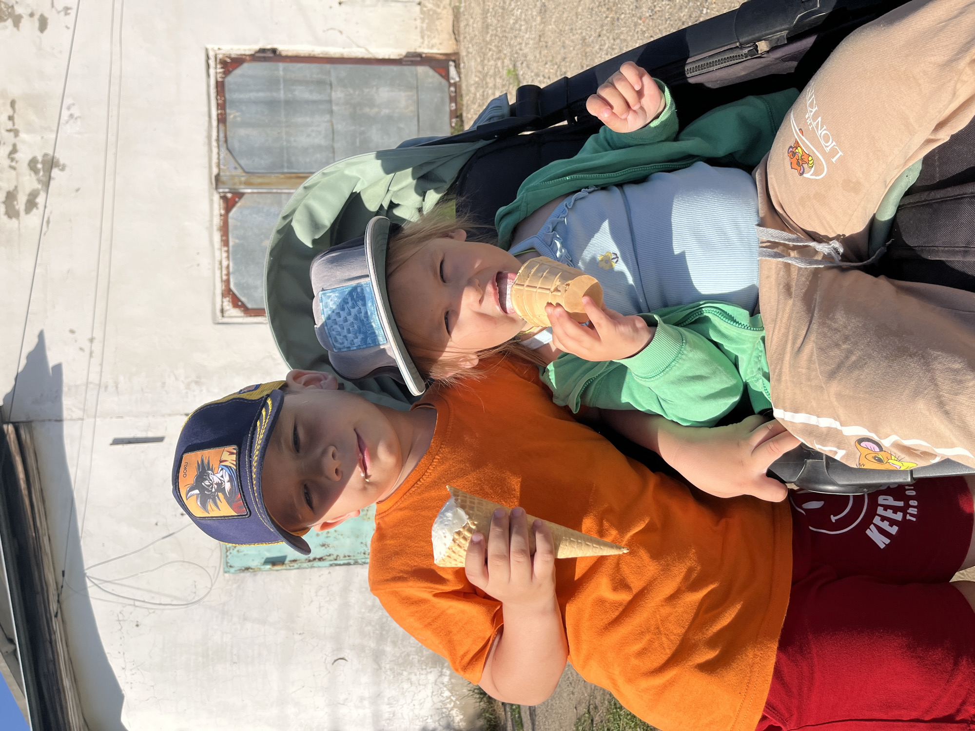 Two young children sit in a stroller outdoors. One child wears an orange shirt, a blue baseball cap, and red shorts that read "KEEP the Original" on the leg. This child holds an ice cream cone. The other child wears a green jacket over a light blue shirt, a blue-grey hat, and tan pants with a "Lion King" graphic. This child is eating an ice cream cone. The background shows a white building with a window. The location is Chaikovsky, Russia.