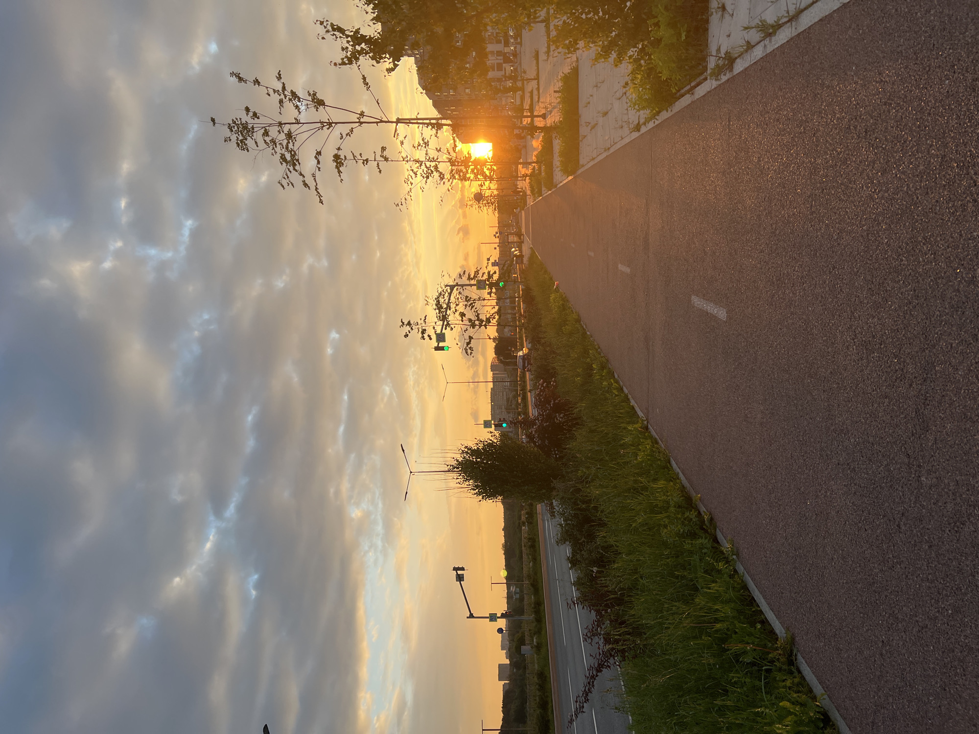 The image depicts a sunset over a city street with a designated bike path. The sun is low on the horizon, casting a warm, golden light across the scene. The sky is filled with dramatic clouds, illuminated by the setting sun. On the left side of the image, a road with traffic lights and distant buildings is visible. On the right, a bike path runs parallel to the road, bordered by grass and trees. The bike path is paved and marked with white lines. The overall atmosphere is serene and picturesque, suggesting a peaceful evening in an urban environment. The scene appears to be a modern development with well-maintained infrastructure. The location is identified as Yekaterinburg, Russia, suggesting it is a public space designed for recreation and transportation within the city.