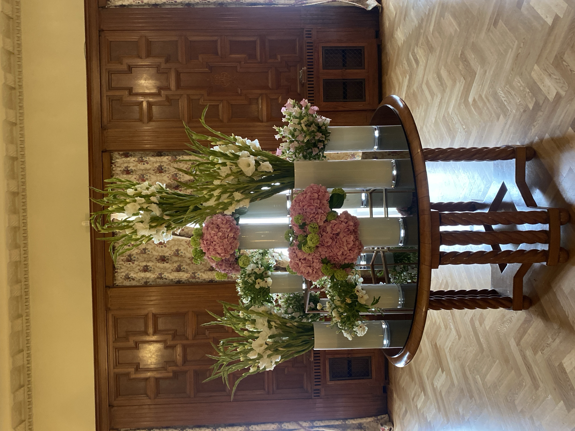 The image depicts a richly decorated room, possibly a grand hall or entryway, featuring a round table laden with floral arrangements. The table has a dark, reflective top and a carved wooden base with twisted legs. The floral arrangements consist of tall white gladiolus, pink hydrangeas, and smaller white and pink flowers in various white cylindrical vases. The background is dominated by dark, ornate wooden paneling and patterned curtains. The flooring is a light-colored parquet in a herringbone pattern. The overall ambiance suggests a luxurious interior, likely within a historic building. The scene does not contain any individuals, nor does it provide any visual cues to pinpoint a specific location within Saint Petersburg, Russia.