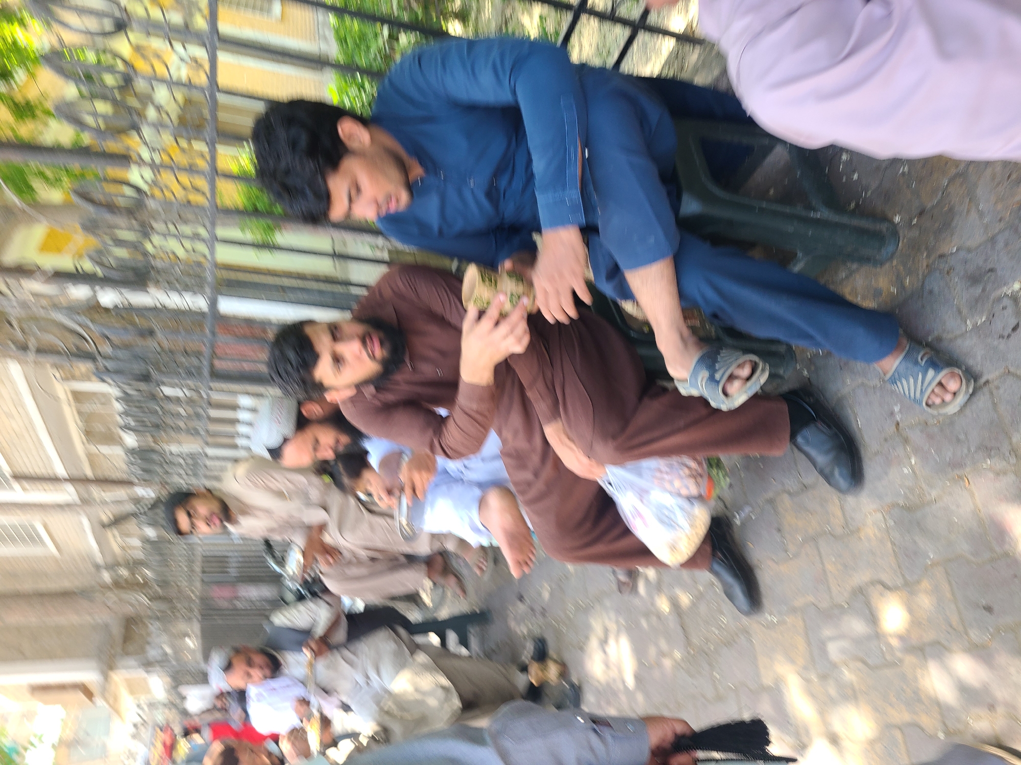A group of men are seated outdoors in Quetta City Tehsil, Pakistan. One man, wearing a blue kurta, sits on a green plastic chair. Another man beside him, in a brown kurta, holds food. Other men are seated on the ground or on low chairs around them, some wearing traditional Pakistani clothing. A metal fence and buildings are visible in the background.