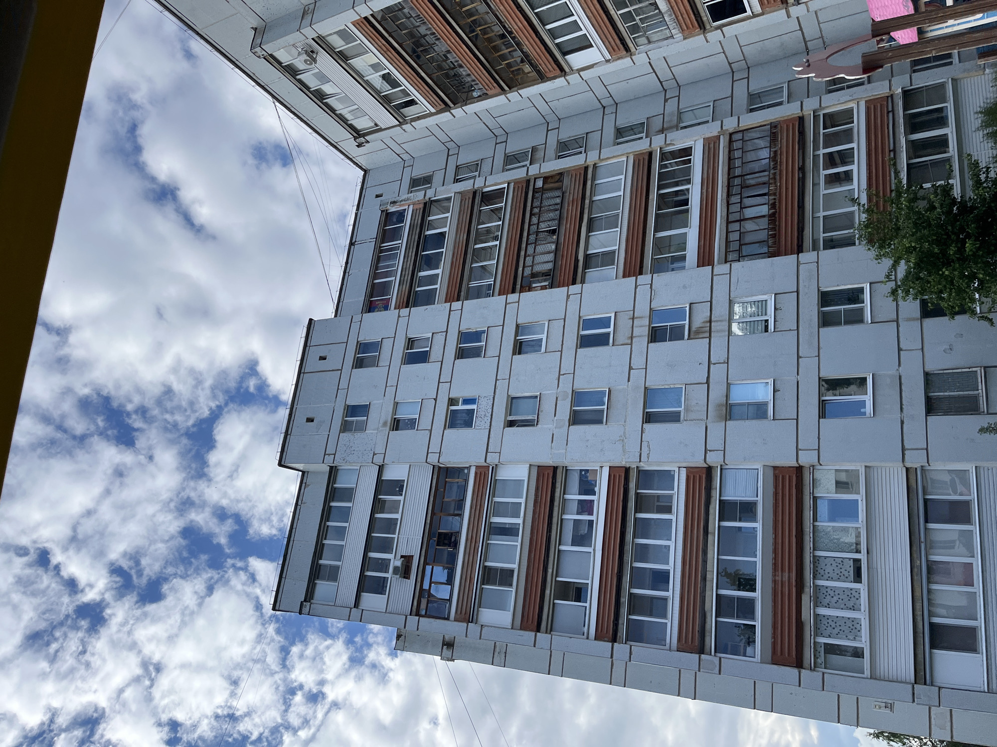 The image presents a low-angle, upward-facing view of a large, multi-story building under a bright blue sky dotted with scattered white clouds. The building, likely located in Chaikovsky, Russia, features a facade primarily composed of light grey or pale blue rectangular panels, arranged in a grid pattern.
Numerous windows of varying shapes and sizes are visible across the building's exterior. Many are tall, narrow, multi-pane windows, often arranged vertically, while others are smaller, individual square or rectangular windows. Some sections of the facade are accented with weathered, reddish-brown corrugated panels, and a few areas show vertical white siding. The building exhibits signs of age and wear, with visible discoloration on some panels and reflections or internal coverings seen through the windowpanes.
A small amount of green foliage, possibly from a tree or bush, is visible on the right side of the structure, partially obscuring a section of the building. On the far left of the frame, a segment of a yellow structure or wall is visible. In the upper portion of the image, a different building or an upper section of the same complex is partially visible against the sky, sharing a similar architectural style. Thin wires or cables stretch across the sky in the mid-ground. The bright, cloudy sky indicates daytime. No discernible text is visible on the building or in the scene.