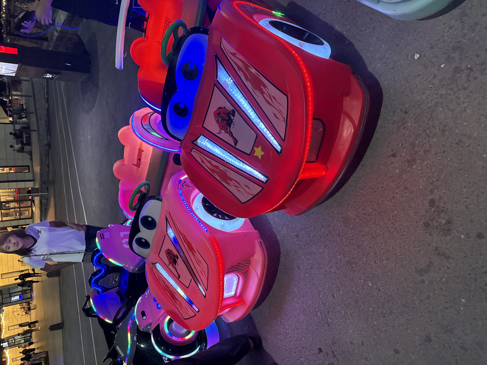 A woman stands behind several illuminated, cartoon-style ride-on cars, likely in an entertainment area. The cars, predominantly pink and red, feature large, expressive eyes and are adorned with LED lights in various colors including blue, pink, and red. Some cars have decals of flames and a bull graphic. The ground appears to be asphalt. In the background, a building with illuminated windows and people can be seen. The lighting suggests it is evening or nighttime. The location is not identifiable as Pavlodar, Kazakhstan, from the provided image.