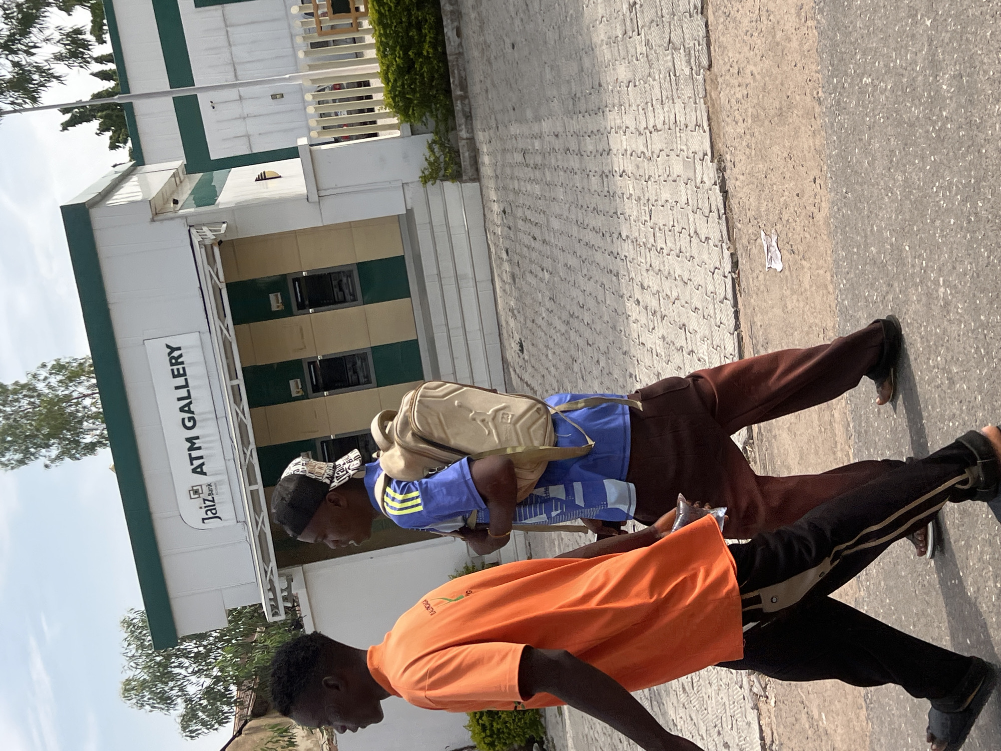 Two young men are walking on a paved sidewalk in Jalingo, Nigeria. The man in the foreground is wearing an orange t-shirt and black pants with stripes. He is walking away from the camera. Behind him, another young man is walking in the same direction. He is wearing a blue t-shirt with yellow stripes on the sleeves and a beige backpack. He also appears to be wearing a patterned head covering.
The background features a white building with green trim. A sign on the building reads "Jaiz Bank ATM GALLERY". The sky is bright, suggesting it is daytime, likely sunny given the shadows. The pavement is a mix of interlocking grey tiles and asphalt. Green foliage is visible on the left side of the building. The overall scene depicts an everyday street view in a Nigerian city. There are no strong indications of emotion; the individuals appear to be going about their daily activities.
