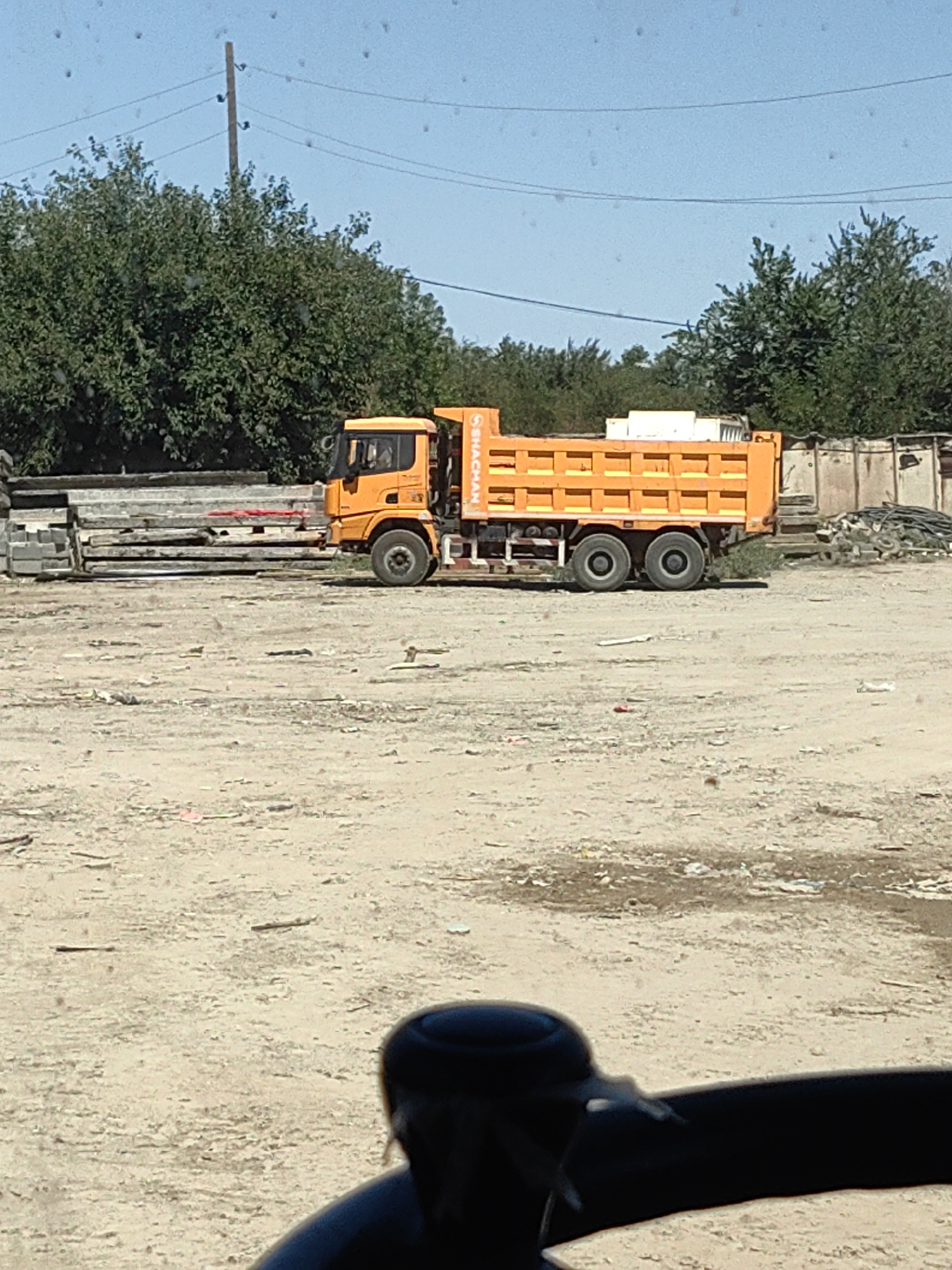 An orange dump truck is parked on a dirt lot. The truck has "SHACMAN" written on the side in white lettering. In the background, there are trees and a few buildings. There are also stacks of concrete slabs to the left of the truck. The sky is blue and clear. The location is Shymkent, Kazakhstan.