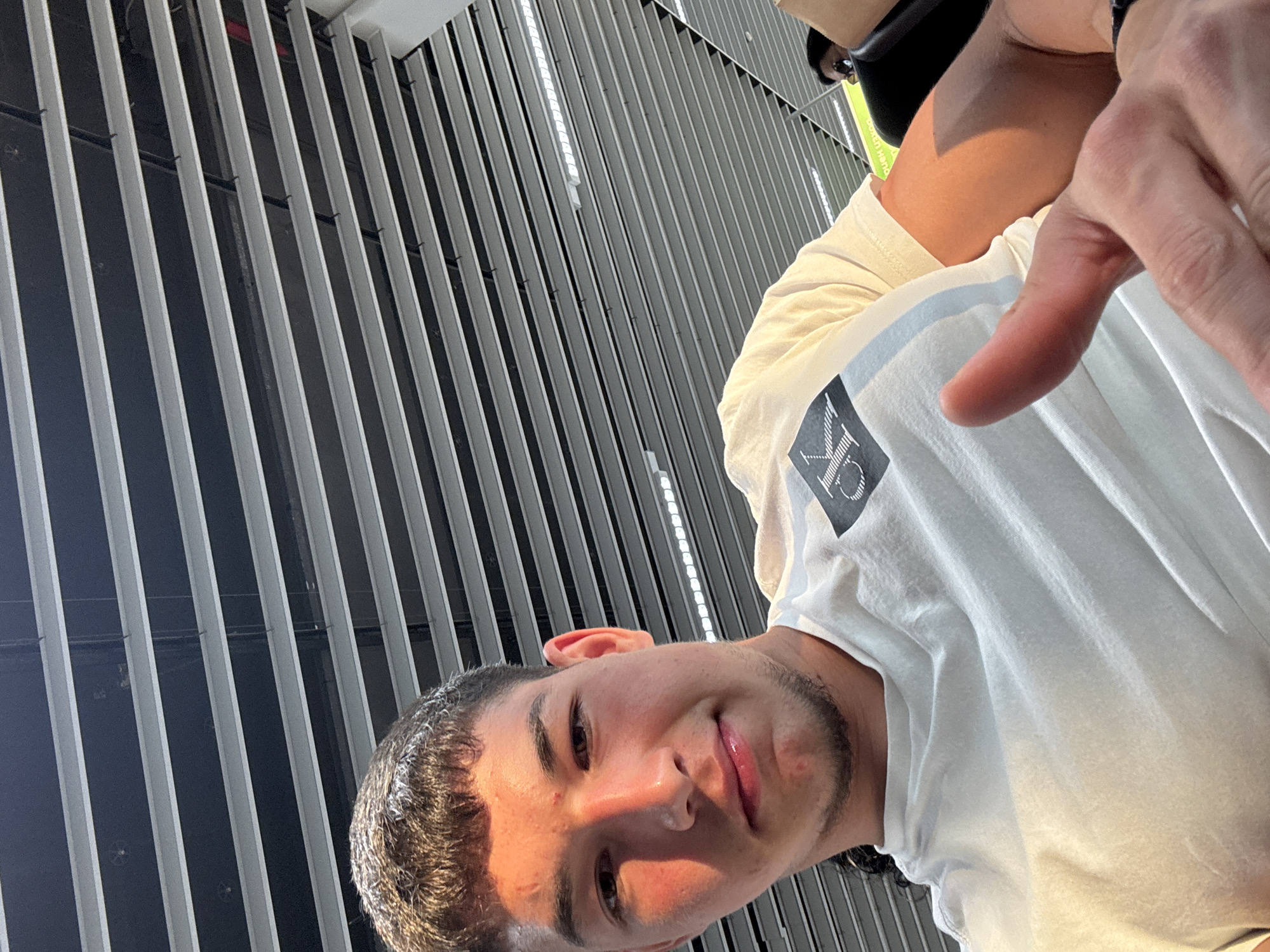 The image is a close-up selfie of a young Caucasian male with short, dark hair and a light beard. He is wearing a white t-shirt with a black logo. His right hand is raised, with his thumb pointing upwards in a "thumbs up" gesture. The subject is looking at the camera with a slight smile. The background consists of a ceiling with parallel horizontal metal beams and some lighting fixtures. The lighting suggests it is daytime, with sunlight casting a warm glow on the subject's face and arm. The location is identified as Sofia, Bulgaria, though no specific landmarks are visible to confirm this. There is a blurred figure of another person in the background, seated.