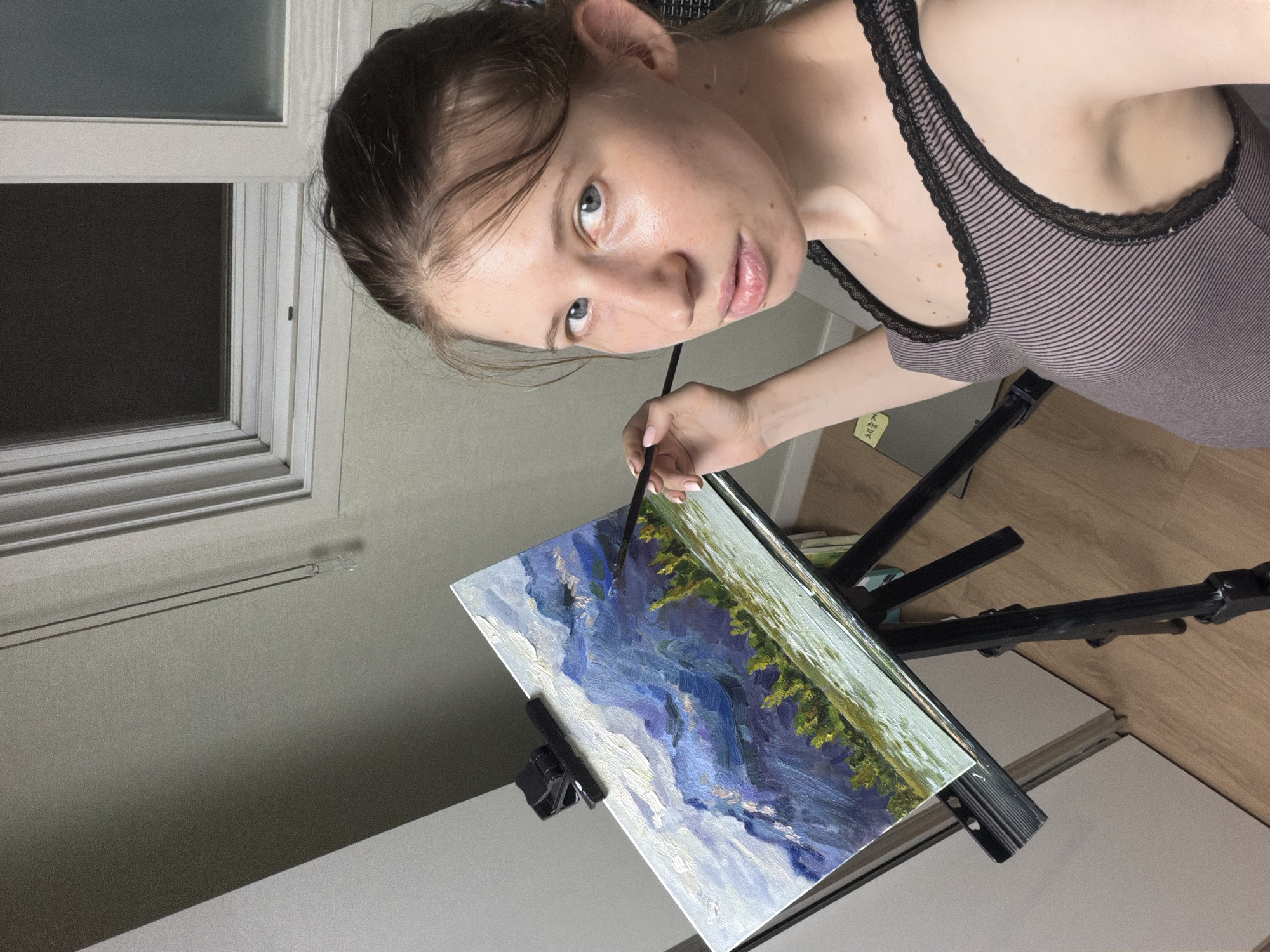 The media file shows a young woman painting on a canvas, which is propped up on an easel. She is a Caucasian woman with light skin, blue eyes, and light brown hair, which is pulled back from her face. She is wearing a striped tank top with black lace trim. Her gaze is directed towards the viewer, and her expression appears neutral, perhaps a bit curious.
The painting on the canvas is an abstract landscape, featuring bold strokes of blue and white for what appears to be water or sky, with touches of green and yellow representing trees or foliage. The painting is still in progress. The woman is holding a paintbrush in her right hand, about to add to the canvas.
The setting appears to be indoors, possibly an art studio or a room in a home. Behind the woman, a window with white blinds is visible, suggesting it might be daytime, though the light is somewhat subdued. The floor is wooden. A black easel supports the painting. There is a hint of what looks like a sticker or label with some Korean text on it, attached to the easel. The overall impression is of a candid moment captured while the artist is engaged in her creative process.