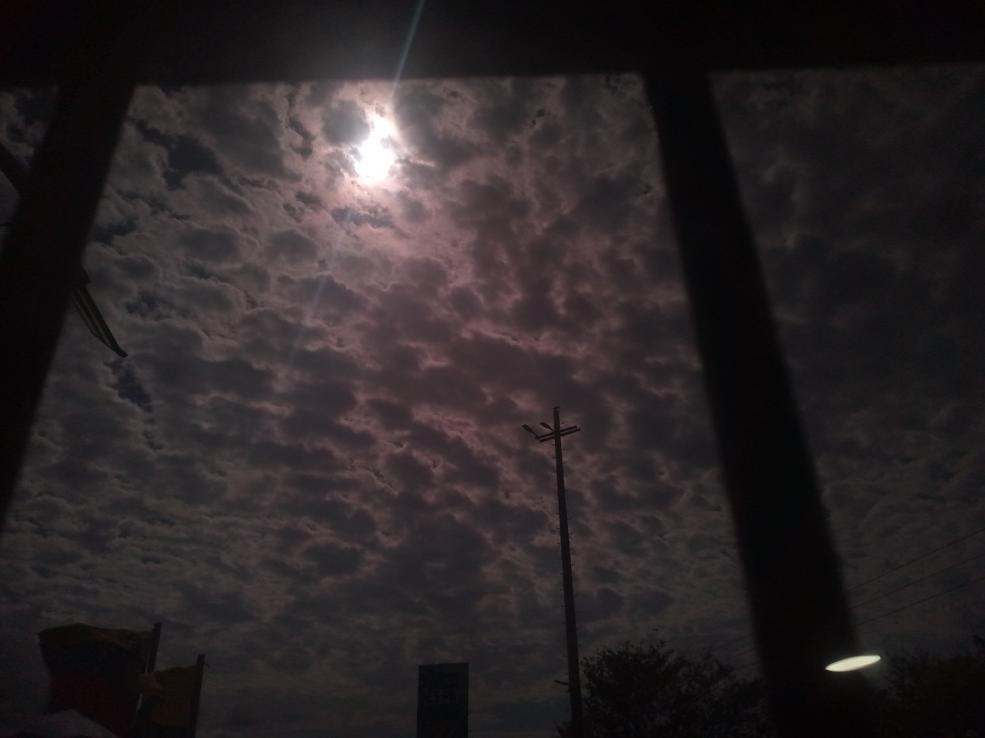 The image captures a view of the sky through a structure, possibly a building or window frame. The sky is filled with clouds and the sun is visible. A utility pole with a streetlight is seen in the distance. In the foreground, flags are visible on the left. The location appears to be an urban environment in the city of Unknown, Unknown.