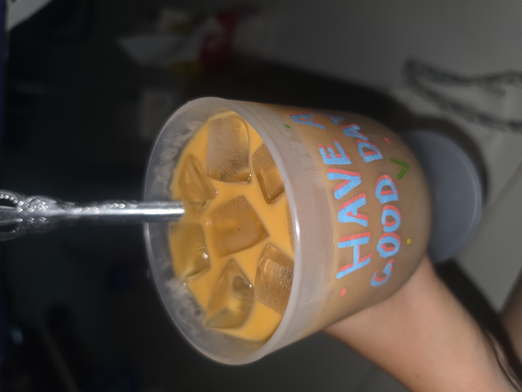 A close-up shot depicts a clear plastic cup filled with an iced orange beverage, likely coffee or tea, with a silver straw inserted. The cup is decorated with the words "HAVE A GOOD DAY" in alternating blue and red colors. The cup is held by a person, their hand visible in the lower right. The background is indistinct, but appears to be indoors. Location is Unknown, Unknown.