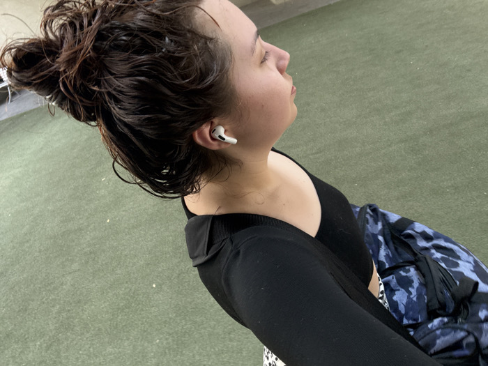 A single individual, an adult female, is depicted in profile from the upper torso, facing the right side of the frame. Her eyes are closed or cast downwards. Her dark brown hair is wet and pulled back into a bun, with some loose strands around her face and neck. A white wireless earbud is visible in her right ear. She is wearing a black tank top or crop top underneath a black long-sleeved top or cardigan, partially visible on her shoulders and arms. To her right, a blue and black camouflage or patterned bag or garment is partially visible. The background consists of a uniform dark green surface, possibly artificial turf or painted concrete, with scattered light-colored debris. The lighting is even. There are no discernible architectural features or landmarks to identify a specific location within Mexico City.