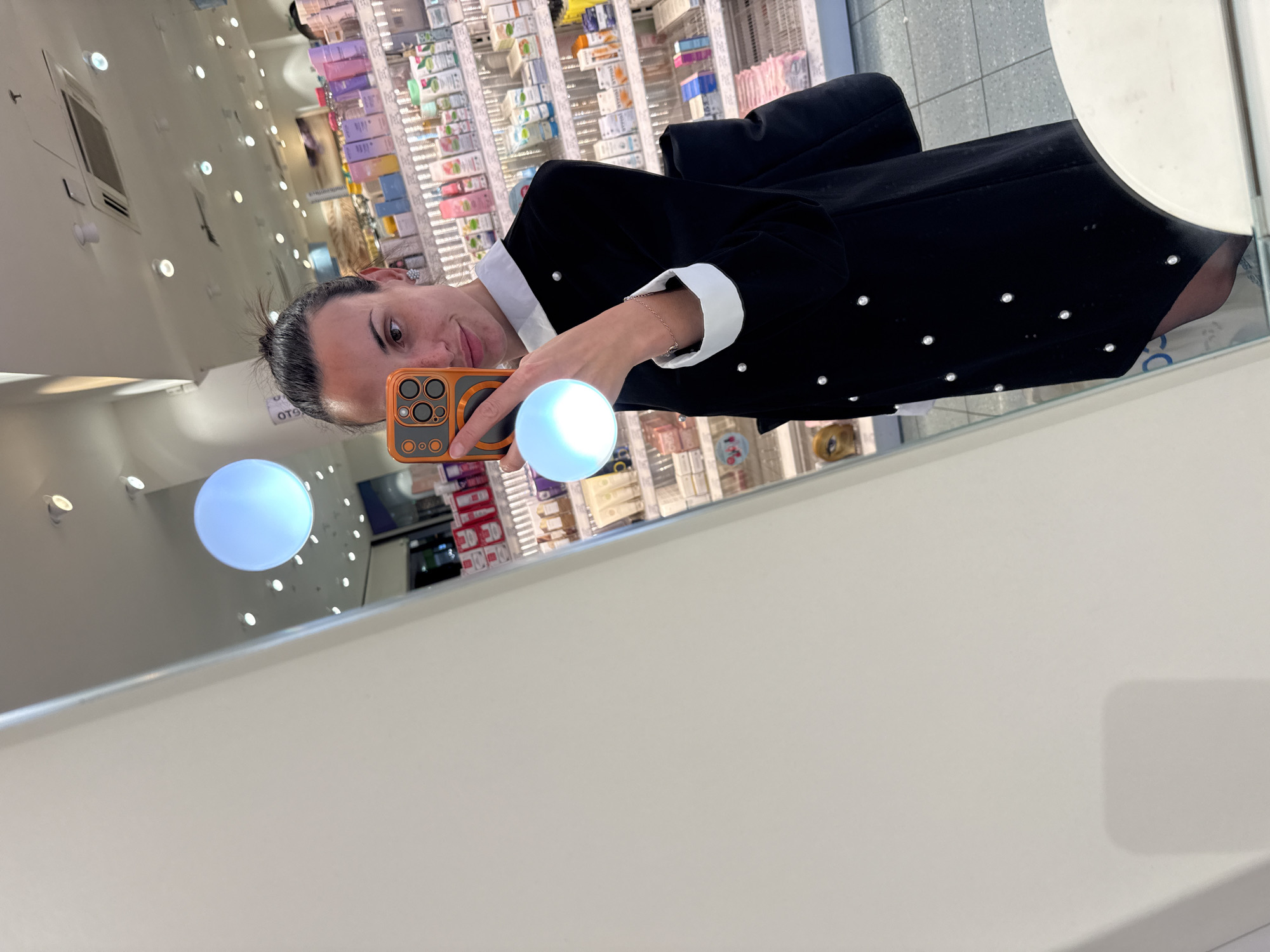The image shows a person taking a selfie in front of a mirror. The reflection shows the person wearing a black dress with white cuffs and decorative pearls. They are holding an orange smartphone. In the background, a shelf stocked with various products is visible. The location appears to be inside a retail store, but there are no explicit indications to pinpoint it within the city of Pleven, Bulgaria.