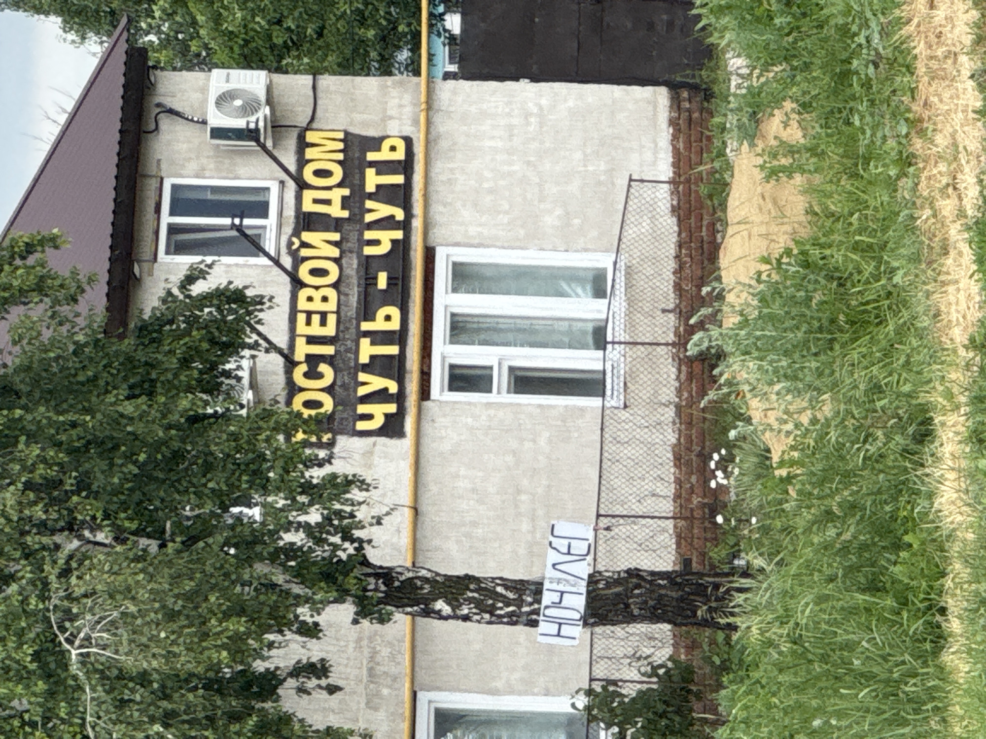 This is a medium shot, slightly high-angle, eye-level photograph of a building with a sign that reads "ПОСТЕВОЙ ДОМ ЧУТЬ-ЧУТЬ" in yellow Cyrillic letters against a dark background, positioned above another line of text. The building appears to be a single-story structure with a dark brown pitched roof. There are several windows visible on the facade, some with white frames. An air conditioning unit is mounted on the exterior wall next to a window. A birch tree with a thin trunk and green foliage stands to the left of the building, partially obscuring it. A white sign with black Cyrillic lettering, reading "НОЧЛЕГ," is attached to the birch tree. A yellow pipe runs horizontally across the building's facade. A chain-link fence with a red brick foundation is visible in front of the building. The foreground is filled with green grass and vegetation. The sky is overcast and appears to be daytime. The scene is located in the Voskresensky District, Russia.