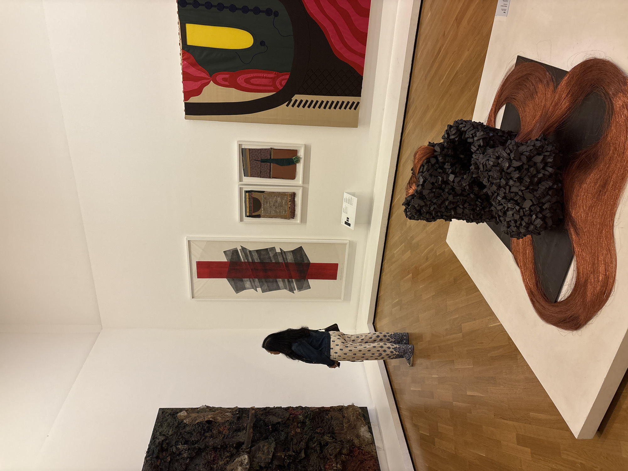 A woman stands in an art gallery in São Paulo, Brazil. She is wearing a dark denim jacket, patterned pants, and sneakers, and faces a framed artwork featuring red and gray geometric shapes. The gallery exhibits a variety of contemporary art pieces, including a large abstract painting with bold colors and geometric patterns, two smaller framed textile pieces, a dark, textured artwork near the floor, and a three-dimensional piece on a pedestal that appears to be constructed from black material and red hair-like strands. The gallery has white walls and a wooden floor.