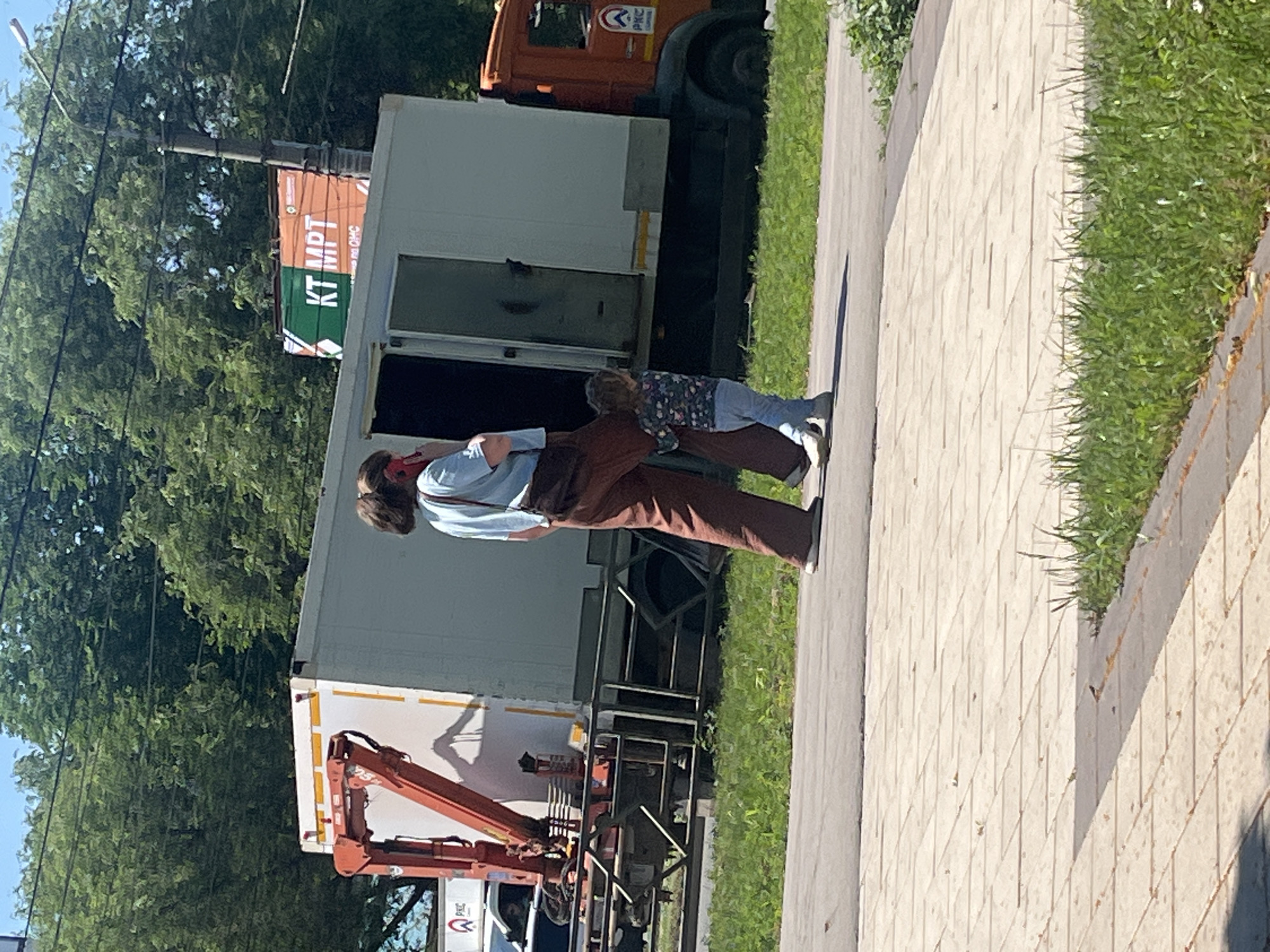 The media file shows a person, likely an adult, interacting with a small child. They are standing on a paved walkway next to a grassy area. Behind them is a large white truck with its doors open, and further back, an orange truck. The scene appears to be outdoors, under a bright sky, suggesting daytime with sunny weather. The person is wearing a light blue shirt and brown pants, and the child is dressed in a colorful patterned top and light blue pants. The adult has their arm around the child, and their posture suggests a close, possibly affectionate, interaction.
A prominent detail is the sign on a pole behind the people and trucks. It has the text "KT MDT" in large letters, with smaller text below. Another visible detail is the side of the white truck, which has a large, dark opening. Part of an orange mechanical arm is visible on the side of the white truck, suggesting it might be some sort of service vehicle or be involved in a delivery or setup. The paved walkway is made of light-colored square tiles. The overall impression is a casual outdoor scene, possibly in an urban or suburban setting, with a moment of connection between the adult and child.