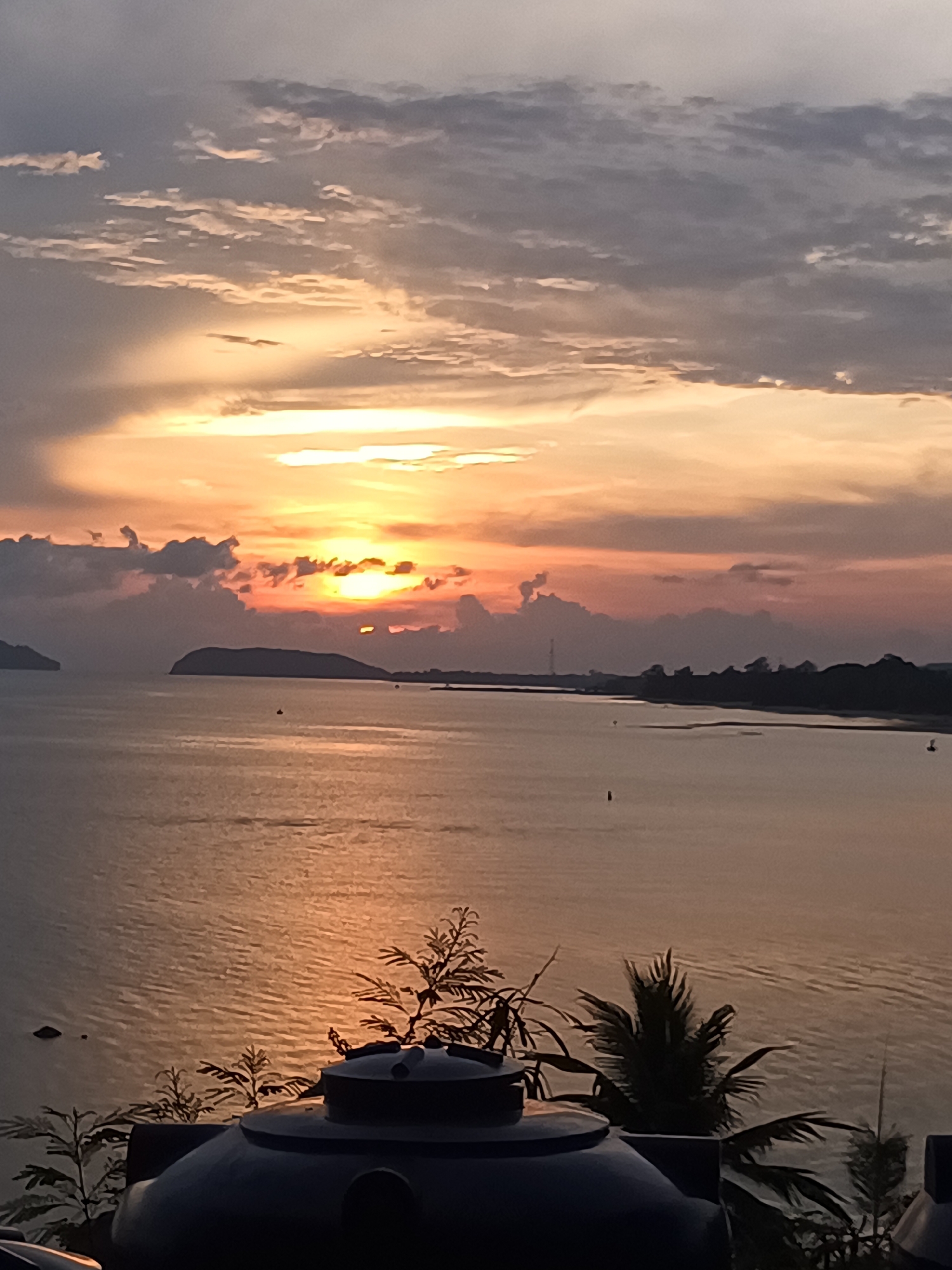 The image captures a sunset over the ocean in Ban Haad Rin, Thailand. The sun is setting behind clouds, casting a golden glow on the water. The coastline is visible in the distance, with a few small islands. In the foreground, there are some plants and a water tank. The sky is filled with clouds, ranging in color from gray to orange. The overall scene is peaceful and serene.