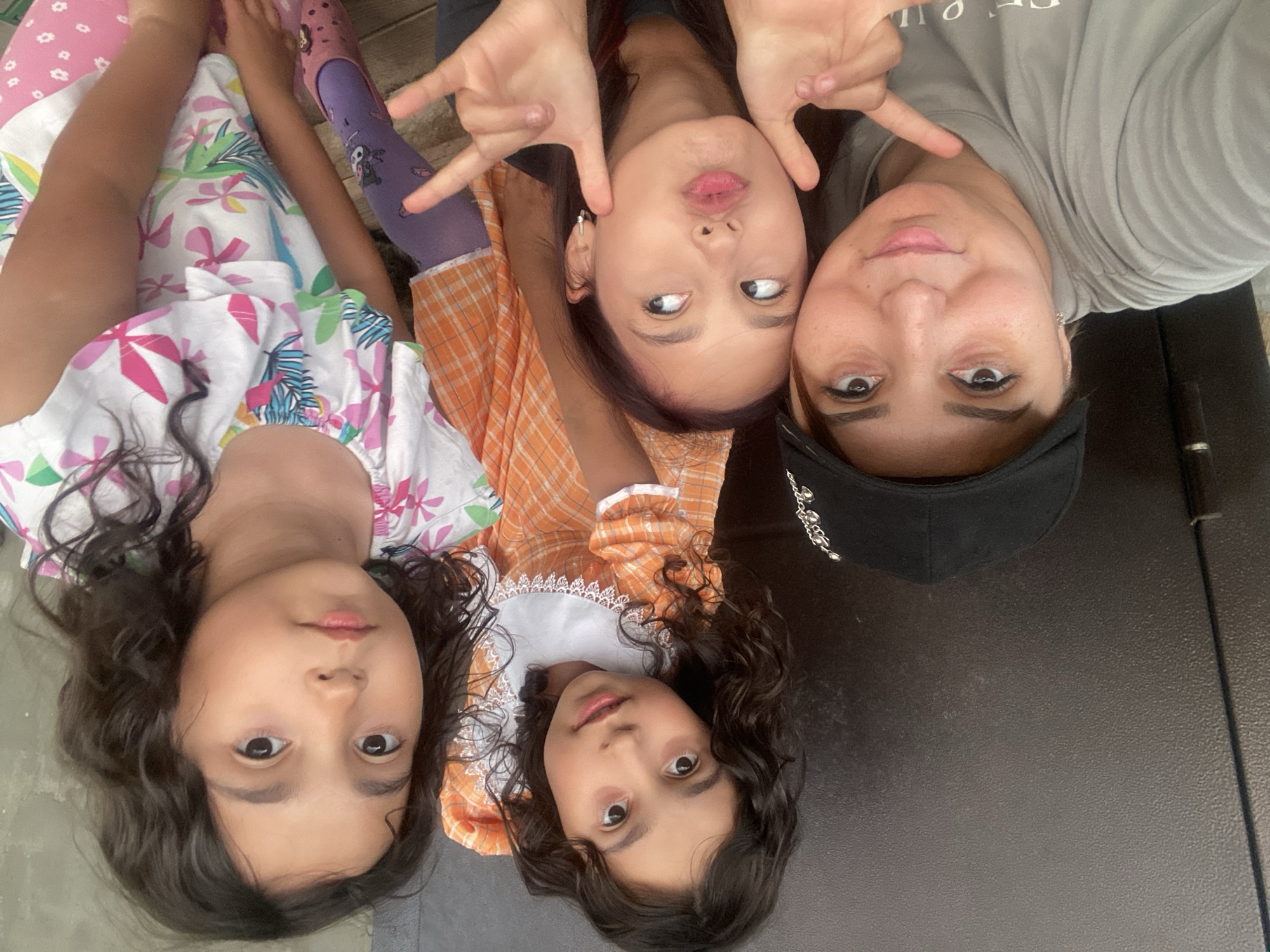 A woman wearing a black baseball cap and a grey t-shirt is taking a selfie with three young girls. The woman is in the foreground on the left, with the three girls behind her. The girl closest to the camera is making a playful face with her hands near her cheeks. Another girl in an orange and white checkered dress is looking towards the camera with a slight smile. The third girl, on the far right, is also looking at the camera. They are outdoors, with a dark, textured wall behind them. The image appears to be taken on a sunny day. The metadata of the image indicates it was taken in the city of Tuymazy, Russia.