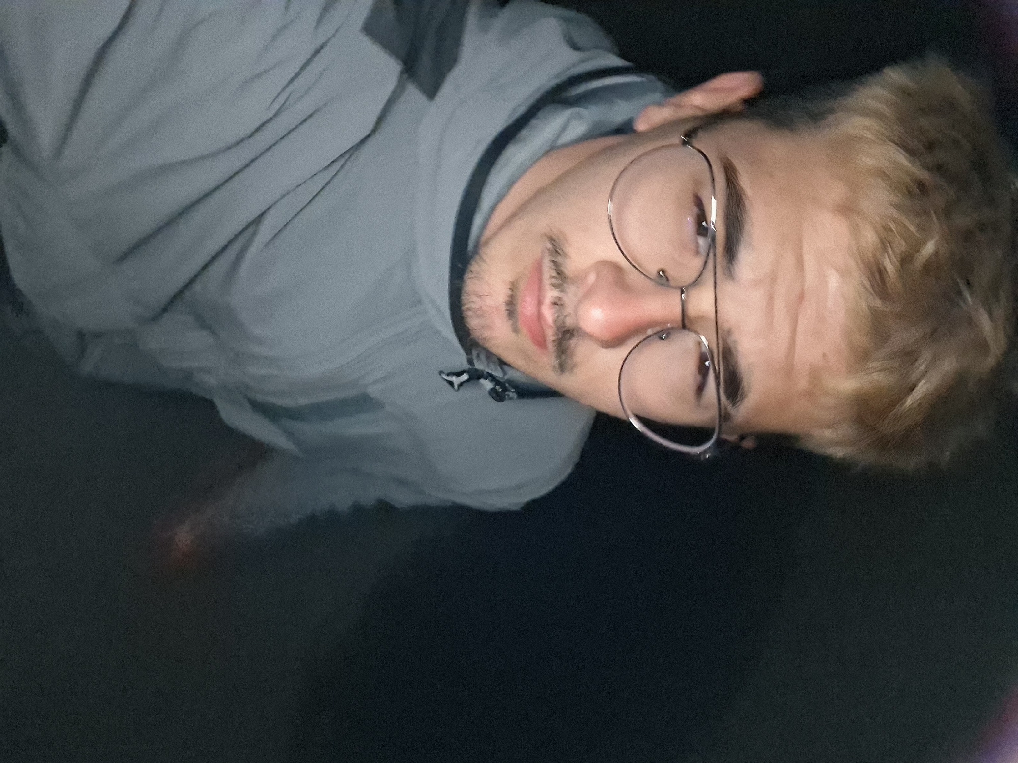 A close-up, slightly angled photo captures a young man with fair, dyed-blonde hair, wearing round, wire-rimmed glasses. He has light skin and a trimmed beard and mustache. He is wearing a gray jacket with black accents on the shoulders. The background is dark and undiscernible. There is no indication of the location being Gabrovo, Bulgaria.