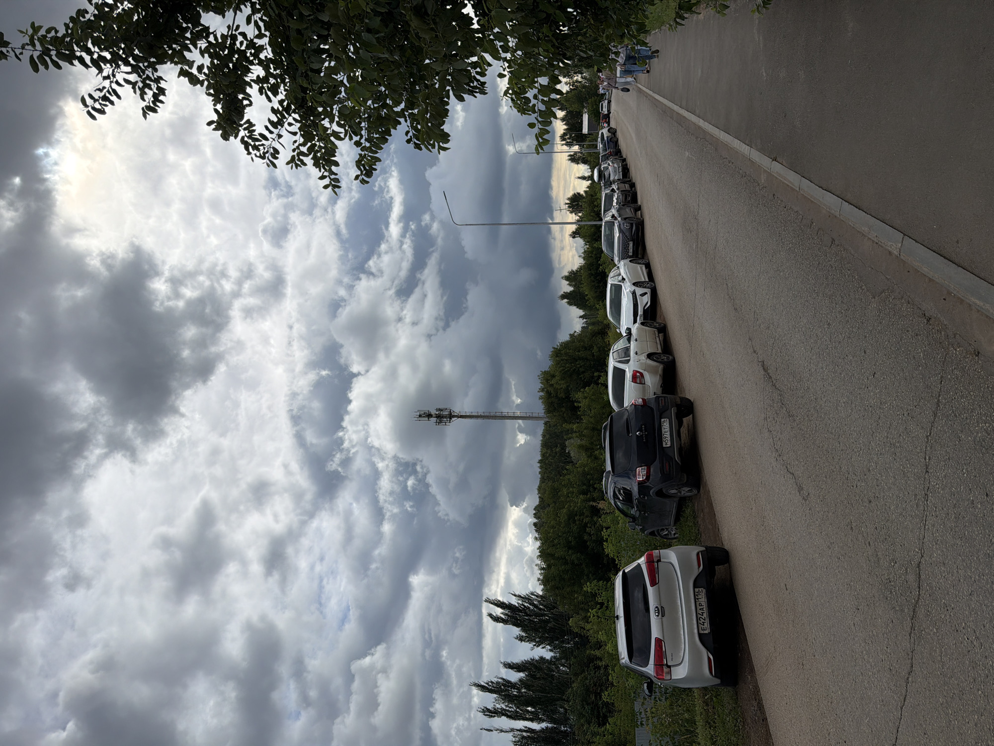 A roadside scene in Yelabuga, Russia, features a line of parked cars along the right side of a two-lane asphalt road. The sky is heavily overcast with dramatic grey clouds. On the left side of the road, a white Kia Rio is parked, followed by a dark SUV with a license plate reading "M597ET 716". Further down the road, several other cars are parked in a single file, including a white Dacia Sandero and a white sedan. A row of tall, slender trees stands behind the parked cars, with denser green foliage beyond them. In the background, a tall telecommunications tower rises above the treeline. On the right side of the road, a paved sidewalk is visible, with a few pedestrians walking away from the camera. A street lamp is positioned along the roadside. The overall environment suggests a rural or suburban setting on a cloudy day.