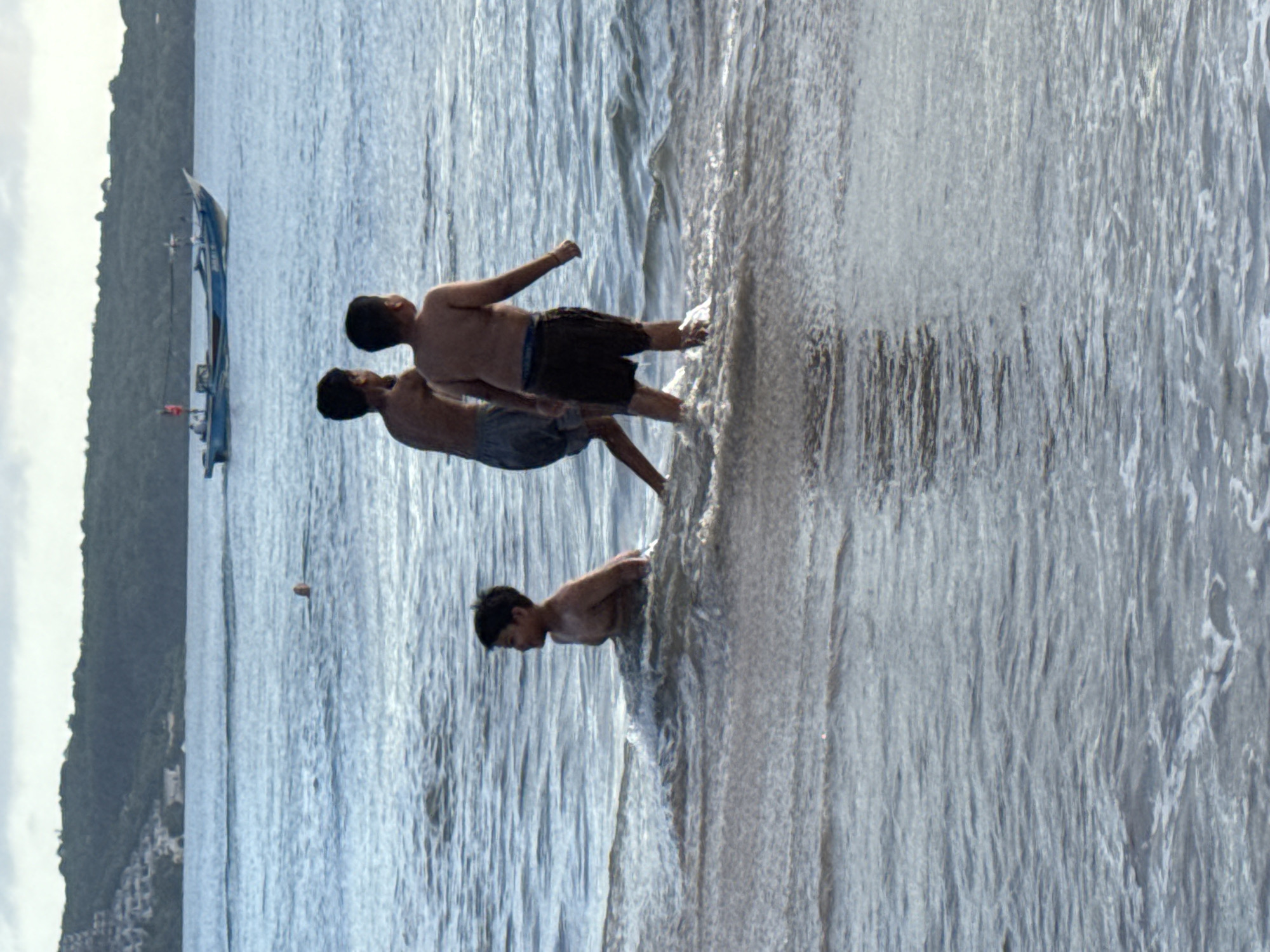 Three shirtless boys are standing at the ocean's edge in Kuta, Indonesia. The first boy, standing closest to the viewer, is positioned in the foreground where waves splash onto the sandy shore. The two other boys stand in a line behind him, near a boat, with varying degrees of submergence in the water. In the background of the image, a lush, green landmass stretches along the horizon, complemented by overcast, light-colored skies.