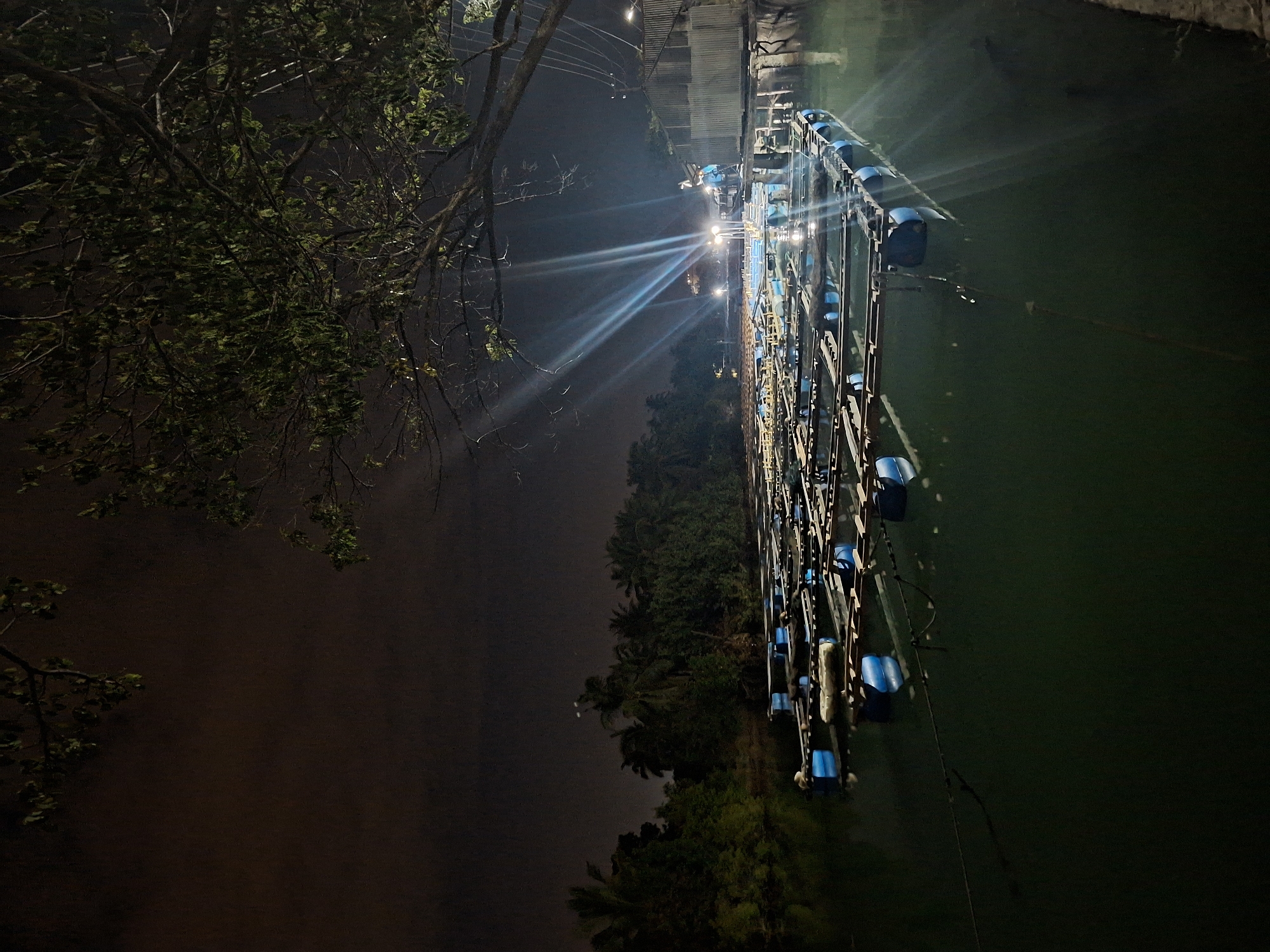 The photograph depicts a nighttime scene in Lhokseumawe, Indonesia. A row of aquaculture equipment, possibly related to shrimp or fish farming, is positioned on the right side of the frame, appearing to be partially submerged in dark water. The equipment features metal frames and blue flotation devices. Bright lights shine from the center, casting beams across the scene. Lush trees and foliage are visible along the shoreline. The left side of the image shows a dark, indistinct area, possibly representing land. A tree branch with leaves is visible in the top left corner.