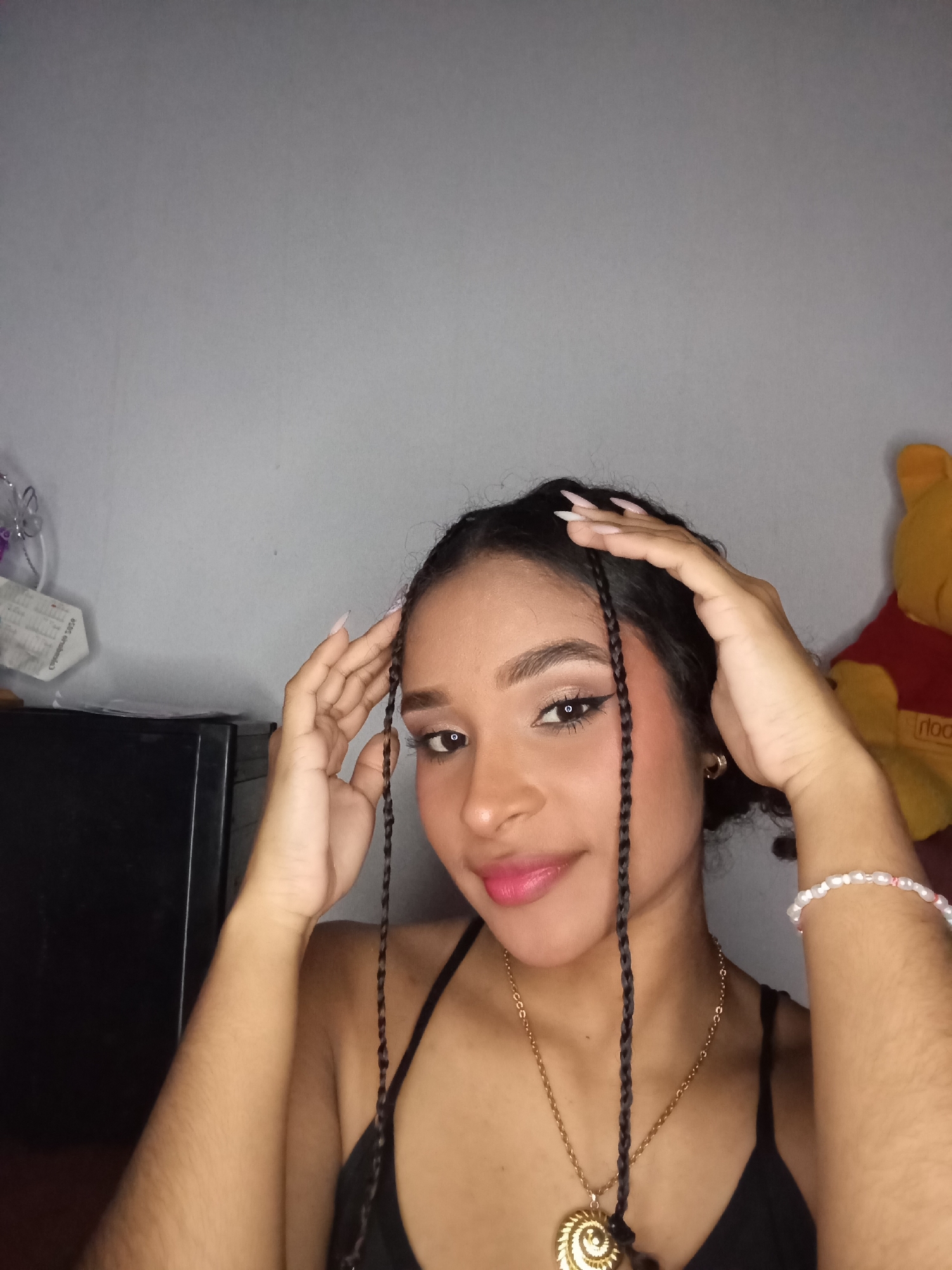 The scene is set indoors, within what appears to be a personal room in the city of Juan Griego, Venezuela. A young female subject is depicted from the chest up, gazing directly at the camera with a slight smile. She has dark, wavy hair, with several thin braids framing her face. Her hands are raised, with fingers touching her hair near her temples. Her fingernails are long, pointed, and painted a light, possibly nude or pale pink color. She is wearing a black top with thin straps. Accessories include gold-colored hoop earrings, a gold-colored necklace with a large, spiral-shell-shaped pendant, and a beaded bracelet with white and pink beads on her right wrist. Her face shows visible makeup, including winged eyeliner, eyeshadow, and pink lipstick. The background features a plain, light grey wall. To the left of the subject, a dark, rectangular object, possibly a small appliance, is partially visible. On this object rests a white item, likely a face mask, bearing text "COVID-19" and "CUIDADO 2020," alongside silver decorative elements. To the right, a yellow stuffed animal resembling Winnie the Pooh, wearing a red shirt with the partially visible text "rloo," is present.