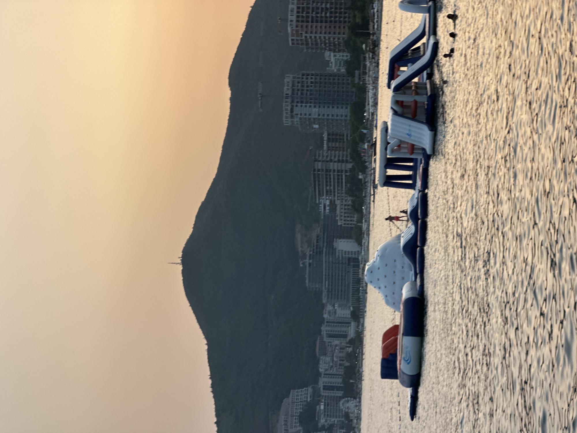 The image shows an inflatable water park floating on the sea in front of the city of Budva, Montenegro. The water park consists of various structures including slides, climbing walls, and a large inflatable iceberg. Several people are visible on and around the water park, with some swimming in the water and others standing on the inflatable structures. In the background, the city of Budva is visible with its buildings and a large, green mountain that dominates the skyline. The sky is a soft, hazy orange, suggesting it is either sunrise or sunset. The sea is relatively calm with gentle ripples reflecting the light. A lone figure is visible on the inflatable iceberg, and several people are swimming in the water to the right of the park. The overall scene depicts a recreational area on the coast of Budva.