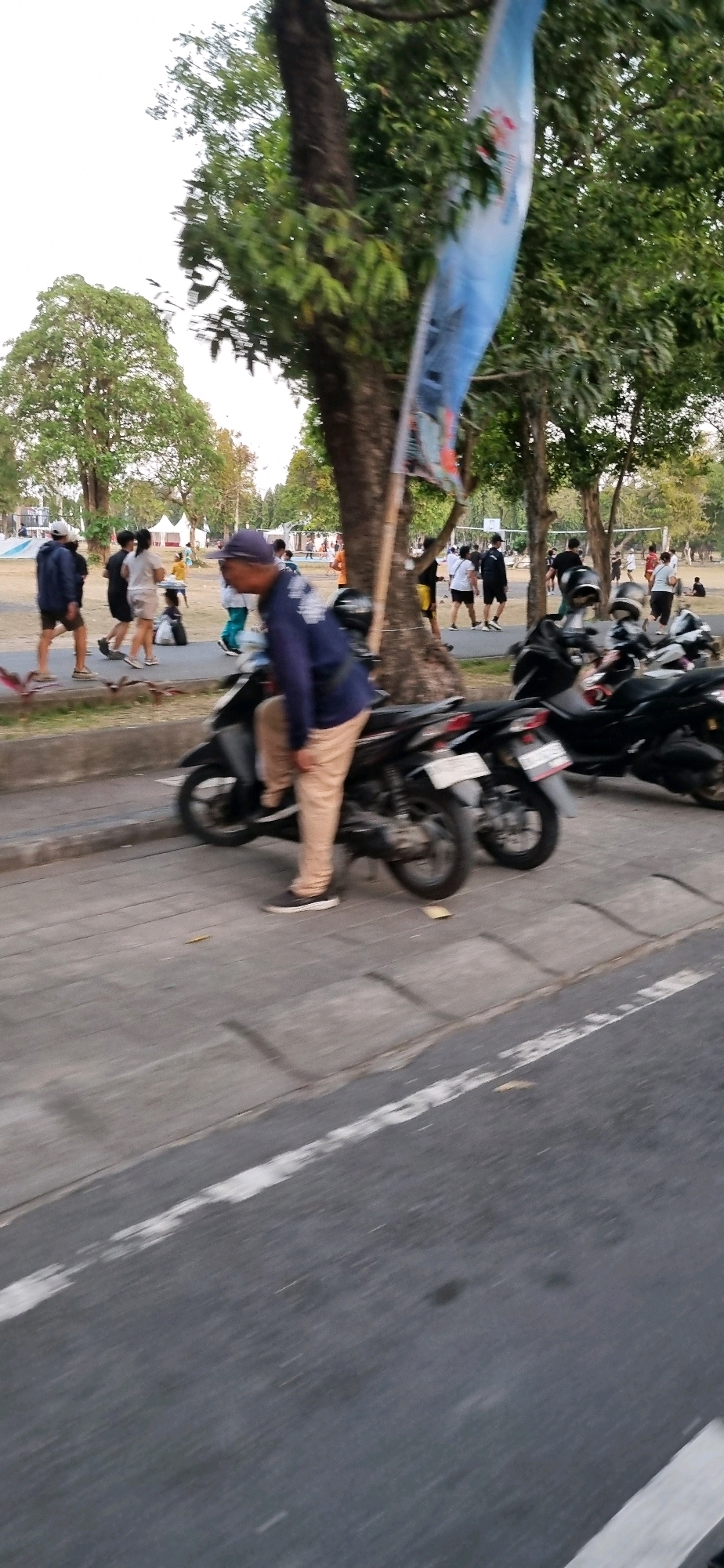 The image captures a street scene in Denpasar, Indonesia. In the foreground, a man wearing a blue long-sleeved shirt and beige pants is leaning over the handlebars of a black motorcycle. He is wearing a dark baseball cap and dark sneakers. The motorcycle is parked on a paved surface, with a sidewalk to its left and a road to its right. Several other black motorcycles are parked in a row behind the first one, with license plates visible on some.
In the background, a group of people are seen walking and standing on a path adjacent to a grassy area. Trees with green foliage are scattered throughout the scene. Some people are dressed casually in t-shirts and shorts. In the far background, there appear to be tents or stalls and a volleyball net, suggesting an outdoor event or recreational area. A blue flag with an image on it is visible, attached to a pole and partially obscured by trees. The overall lighting suggests it is daytime, possibly late afternoon. The image has a slight motion blur, indicating movement during capture.