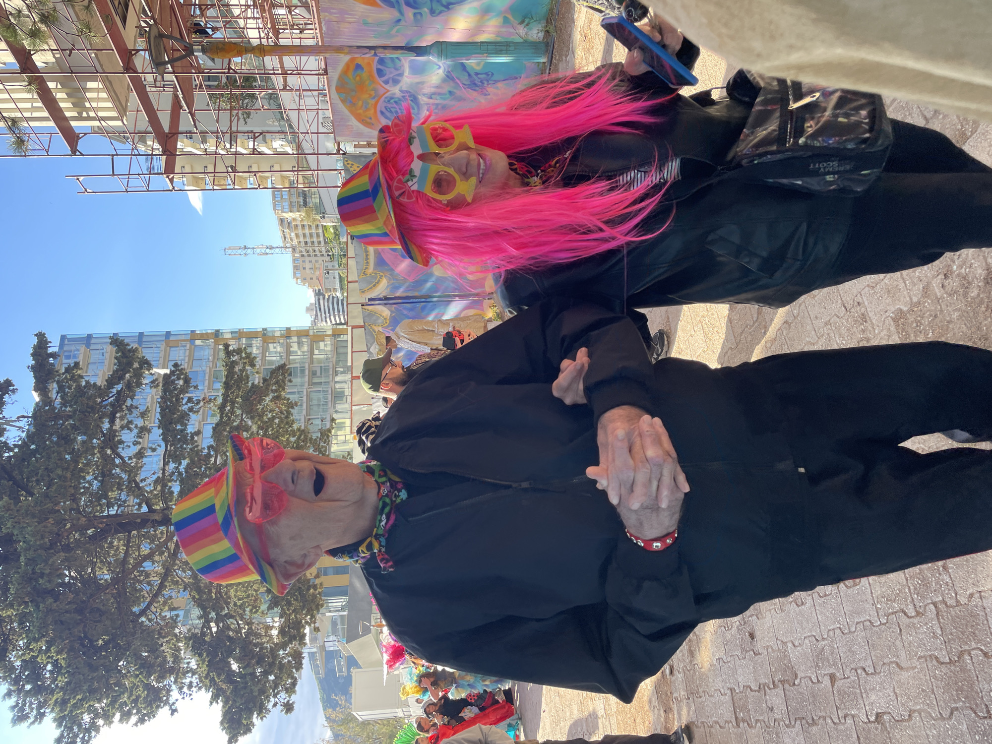 This vibrant outdoor image captures two individuals dressed in festive attire, likely participating in a parade or celebration in Rafailovići, Montenegro.
In the foreground, an elderly person, possibly a man, is prominently featured. He wears a black zipped jacket and black pants, accessorized with a rainbow-striped bucket hat and bright pink heart-shaped sunglasses. A colorful, patterned scarf is tied around his neck, and a red studded wristband is visible on his left wrist. His hands are clasped in front, and his mouth is open, suggesting he is smiling, laughing, or speaking.
Behind him, a second person with striking, spiky pink hair also sports a rainbow-striped bucket hat and yellow novelty glasses. This individual is dressed in a black jacket and holds a blue smartphone in their left hand, smiling broadly. A clear fanny pack, with the text "JEREMY SCOTT" clearly visible, is worn across their body.
The background shows modern buildings with glass facades and some residential structures under a clear blue sky. Sparse trees and what appears to be construction scaffolding or metal frameworks are also visible. Behind the pink-haired individual, a colorful, abstract mural or painted backdrop adds to the festive atmosphere. The ground is paved with light-colored, interlocking bricks. The bright daylight and sunny weather suggest the event is taking place during the day, conveying a joyful and celebratory mood.