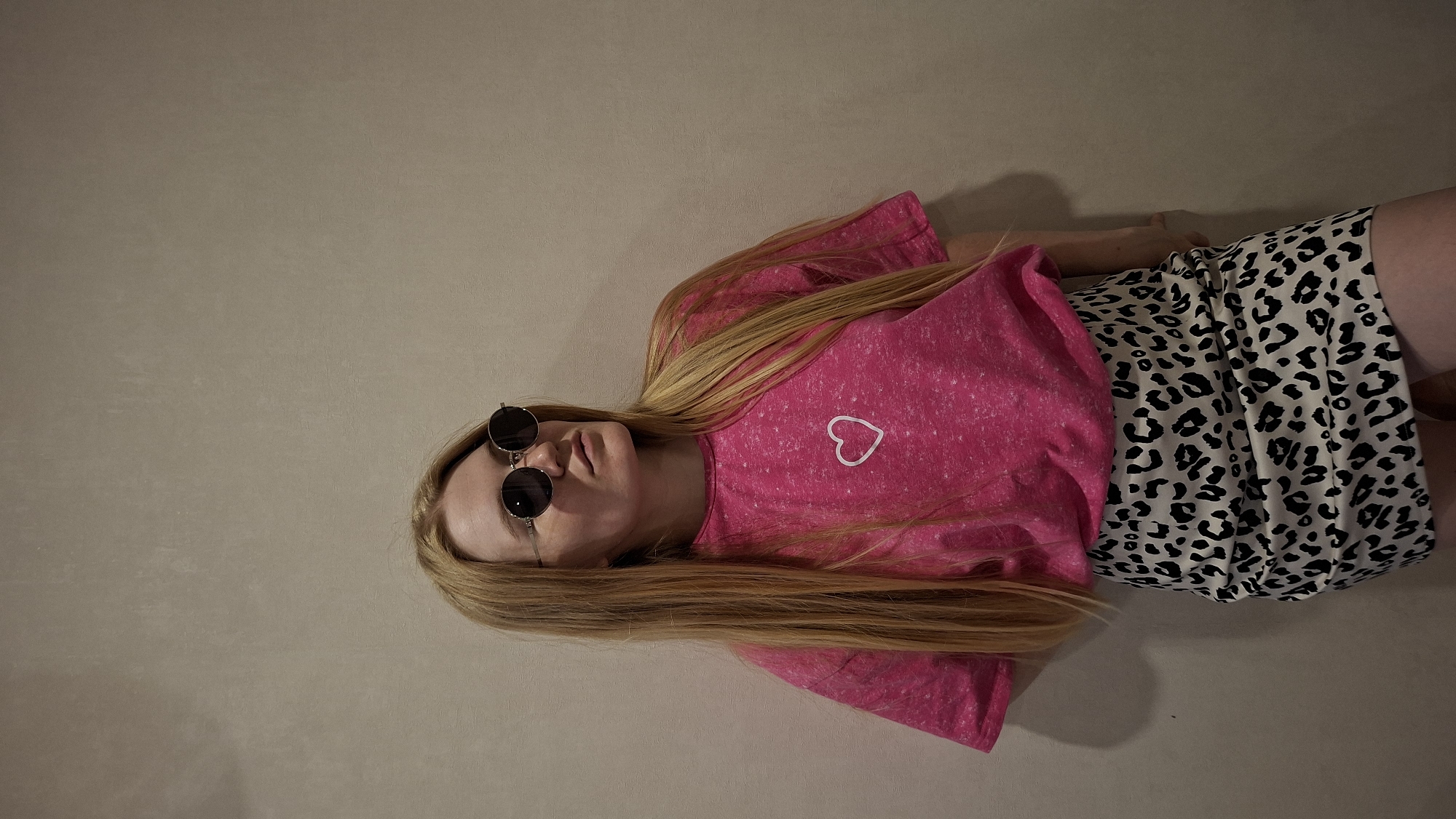 A young woman with long blonde hair is lying on her back on a neutral-colored surface. She is wearing a bright pink t-shirt with a white outline of a heart on the left side of her chest, and a skirt with a black and white leopard print pattern. She is also wearing round, dark sunglasses. The image is taken from a high angle, looking down at the woman. The lighting is soft and diffused, casting a gentle shadow to the right of the woman. The background is a plain, textured wall, suggesting an indoor setting. The overall mood is relaxed and casual. There is no visible text in the image.