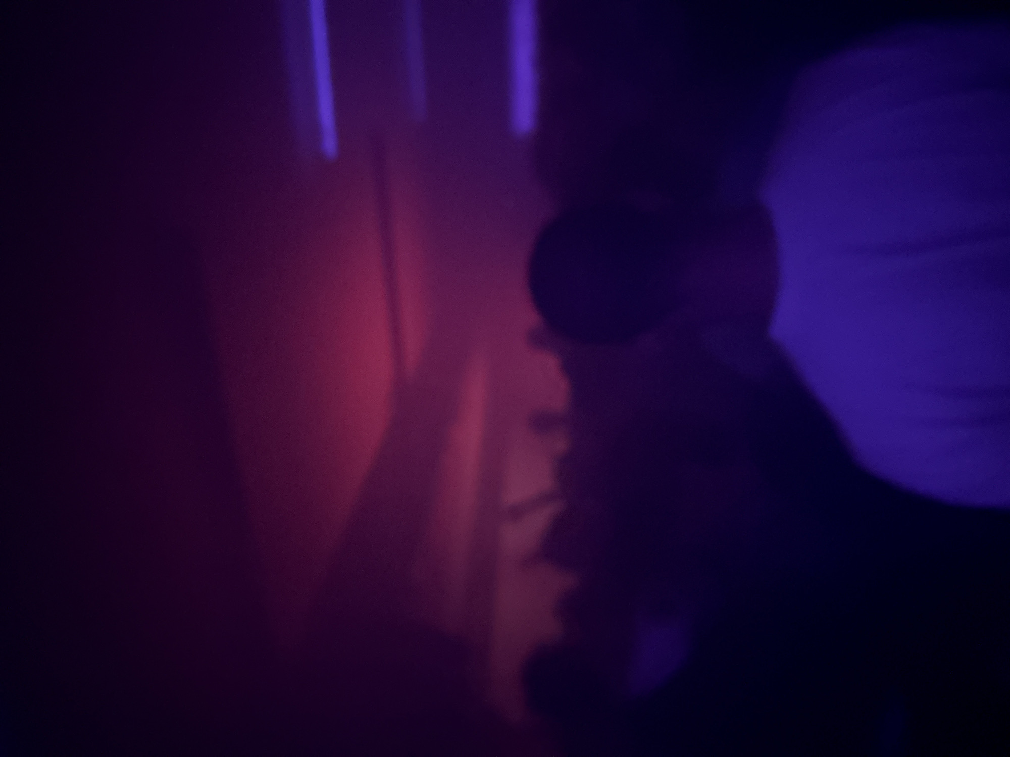 The image shows a person's face in shadow and a nearby wall with vertical light streaks. The lighting appears to be purple and red. There is not sufficient information to determine the location in Paris, France.