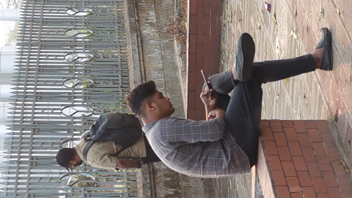 The scene depicts an outdoor urban environment near the Supreme Court of Bangladesh in Dhaka. In the foreground, a young male is seated on a low, red-brick ledge, positioned right of center. He is wearing a grey plaid long-sleeved shirt, dark trousers, and black loafers. His head is bowed, and he is holding a red-edged smartphone with both hands, appearing to interact with it. His left leg is crossed over his right knee. To the left and slightly behind him, a second male figure is partially visible from the back, wearing a light brown shirt, a black baseball cap, and a black backpack. He appears to be standing or walking towards the left. A tall, grey metal fence with vertical bars, pointed finials, and distinctive circular 'Y' shaped decorative elements runs across the mid-ground, separating the foreground from a light-colored building with vertical architectural features in the background. The ground is paved with light-colored tiles, exhibiting scattered dry leaves and a purple wrapper. A weathered grey concrete wall is present behind the brick ledge. The scene is brightly lit by daylight.