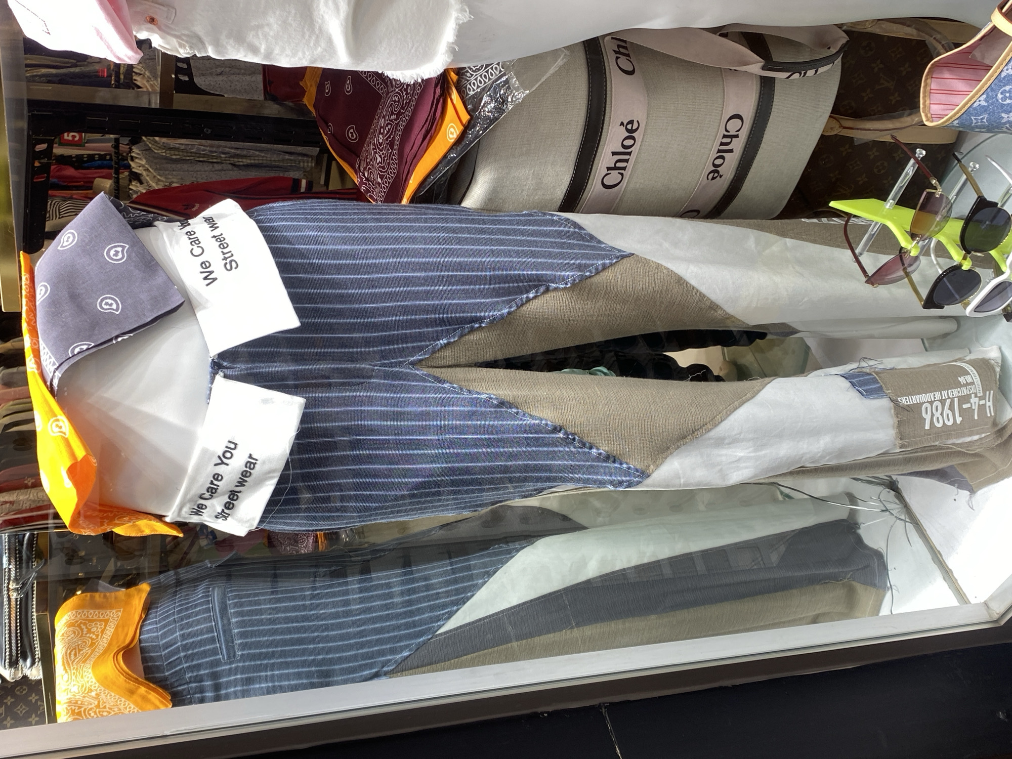 The image displays a collection of clothing and accessories arranged within what appears to be a glass-fronted display case or on shelves in a retail environment, likely in Alanya, Turkey. The central item is a pair of distinctive patchwork pants, constructed from different fabric types: blue pinstriped material, solid beige sections, and plain white fabric.
Adjacent to the pants are folded bandanas in various colors, including grey, bright yellow, and maroon, many featuring a classic paisley pattern. Above these items, a large beige tote bag is visible, prominently featuring "Chloé" branding repeated along its strap. Next to the Chloé bag, another bag with a brown monogram pattern, styled similarly to Louis Vuitton, can be seen. Several pairs of sunglasses with different frame designs and lens tints are arranged on a bright green plastic stand. In the background and around the main items, other garments such as white shirts and folded clothes are visible on shelves.
Visible text on white fabric labels attached to the pants reads: "We Care You Street wear," appearing twice. Another label, brown in color, displays text including "DISPATCHED AT HEADQUARTERS," "H-4-1986," "NO.34," and "INDUSTRIA." The brand name "Chloé" is clearly visible multiple times on the beige bag's strap. Other numerical text, potentially from fabric patterns or background elements, includes "9000," "100," "10000000," and a single digit "5." The scene captures a variety of streetwear and luxury-inspired accessories presented for sale or viewing.