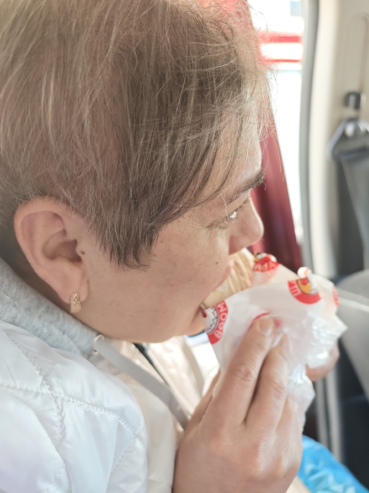 The image depicts an adult individual, observed in right profile from the mid-chest up, consuming food inside a location in the city of Chaikovsky, Russia. The person has short, light brown and grey hair, fair skin with visible freckles, and wears a gold-colored, rectangular, textured hoop earring in their right ear. They are dressed in a light grey hooded sweatshirt beneath a white quilted jacket. The individual holds a food item wrapped in white packaging with red and yellow branding, featuring the word "BOOM" (and Cyrillic "БУМ") and a chicken logo, near their mouth, appearing to eat. The blurred background indicates an indoor setting, with a bright window or opening to the right revealing faint red and white vertical patterns, possibly an exterior view or part of a vehicle's interior. Darker forms on the far right suggest additional interior elements or seating.