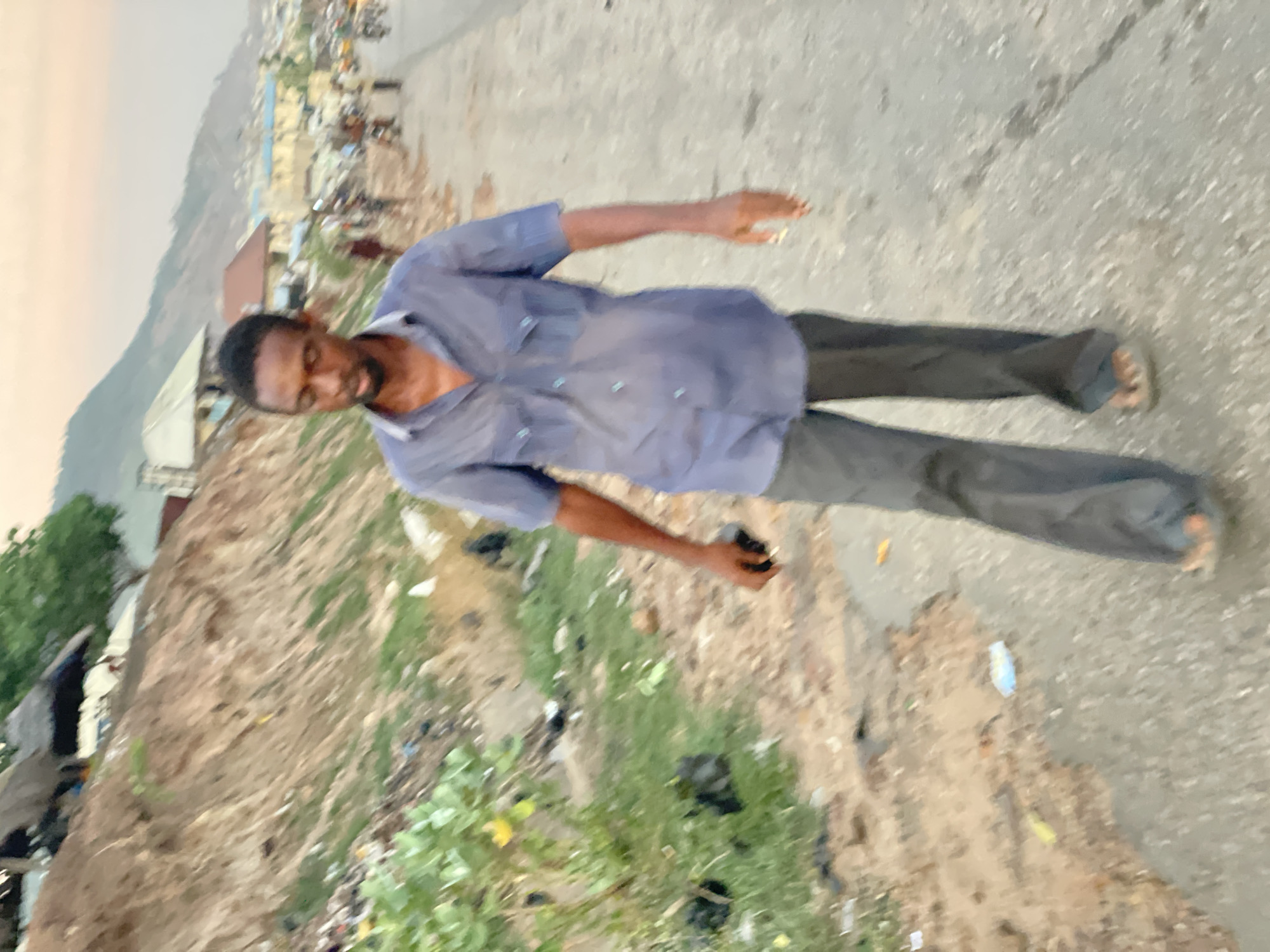 A man stands in an outdoor setting in Lokoja, Nigeria. He has dark skin, short dark hair, and a beard. He is wearing a short-sleeved, buttoned shirt and dark trousers, with sandals on his feet. His arms are extended to the sides. The background shows a mix of urban and natural elements. There's a dirt area containing litter, and a paved road or pathway. In the distance, there are buildings and hills.