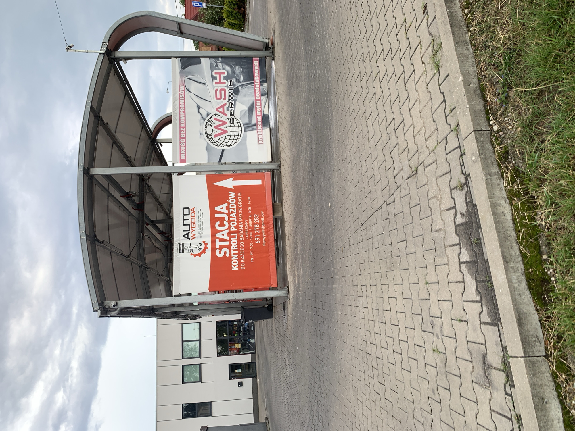 A concrete, paved area with interlocking pavers and a concrete curb is visible in the foreground, with some sparse grass and weeds growing along the curb. In the middle ground, there is a covered structure with a curved roof, supported by metal poles. Two large banners are displayed under the roof. The banner on the left is red and orange and has text in Polish that says "AUTO WYGODA" and "STACJA KONTROLI POJAZDÓW" (Vehicle Inspection Station). The banner on the right is white and features an image of a car and text that says "JAKOŚĆ BEZ KOMPROMISÓW" (Quality without compromise) and "WASH SERWIS" (Wash Service), with "PRODUCENT MYJNI BEZDOTYKOWYCH" (Manufacturer of touchless car washes) below. Behind the covered structure, a modern, light-colored building with large windows is visible on the left. The sky is overcast with clouds. The location is identified as a car wash and vehicle inspection station in Białystok, Poland.