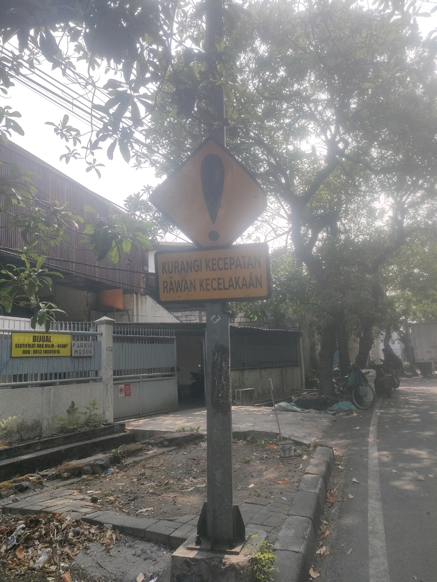 The image depicts a street scene in Surabaya, Indonesia, with a prominent yellow warning sign on a pole in the foreground. The sign is diamond-shaped with a black exclamation mark inside a circle and has text below it. The text on the sign reads "KURANGI KECEPATAN" which translates to "REDUCE SPEED", and "RAWAN KECELAKAAN" which translates to "ACCIDENT PRONE". To the left of the sign, there is a yellow rectangular sign attached to a fence that reads "DIJUAL SERTIFIKAT HAK MILIK - 460M2" meaning "FOR SALE LAND CERTIFICATE - 460SQM" and "HUB: 0813 10005616". Below this it says "PARKIR MASUK" which means "ENTRANCE PARKING". The background shows a building with a corrugated metal roof, a fence, trees with dense foliage, and several motorcycles parked along the side of the road. The road has a white line marking the lane. The ground is a mix of pavement, dirt, and fallen leaves, with some debris visible. The lighting suggests it is daytime, with some haze or sunlight glare obscuring parts of the scene.