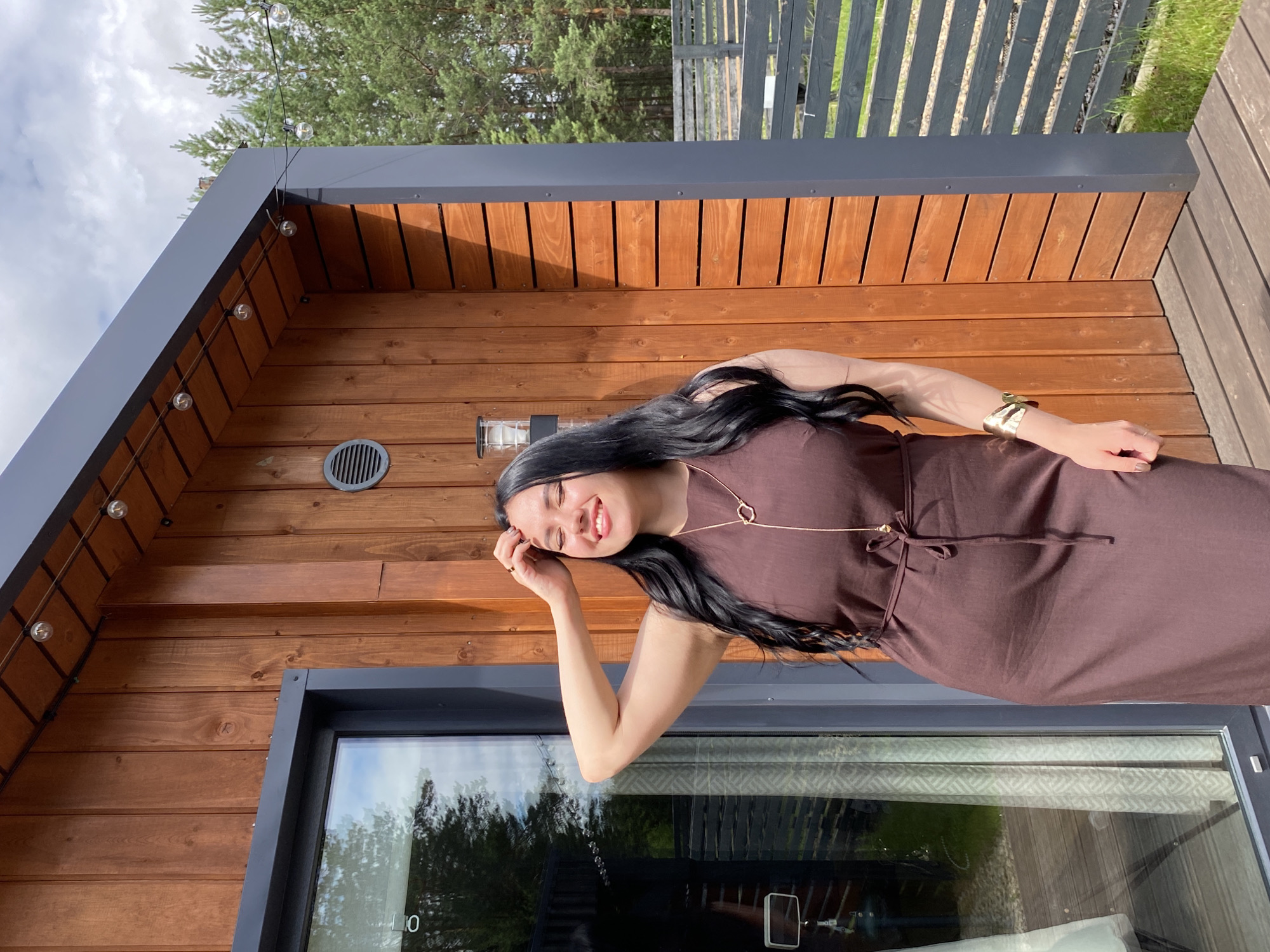 A woman with long, dark hair and closed eyes is standing in front of a wooden building. She is wearing a dark brown dress and a gold necklace. Her right arm is raised, and her hand is resting on her head. The building appears to be a modern, rustic structure with wood siding and large windows. There are string lights draped across the top of the building. In the background, there are trees and a fence, suggesting an outdoor location, possibly in a rural or semi-rural area. The scene is illuminated by natural daylight. The woman's expression appears to be one of contentment or enjoyment. Based on the architectural style and natural surroundings, this location could be a cottage or a modern home in a wooded area, potentially located in or near Chaikovsky, Russia, given the context.