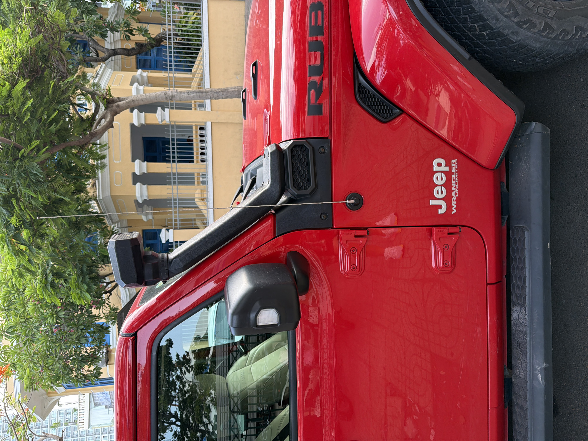 This is a medium shot of the side of a red Jeep Wrangler Unlimited, parked on a street in Nha Trang, Vietnam. The Jeep is facing left, with its front half visible. The vehicle has a black snorkel attached to the A-pillar, a black side mirror with an integrated turn signal, and a black side step. The "RUBICON" and "Jeep WRANGLER UNLIMITED" emblems are visible on the body of the car. Behind the Jeep, a yellow building with white columns and blue shutters can be seen, partially obscured by lush green trees. The ground is paved and appears to be a street. There are no people visible in the image.