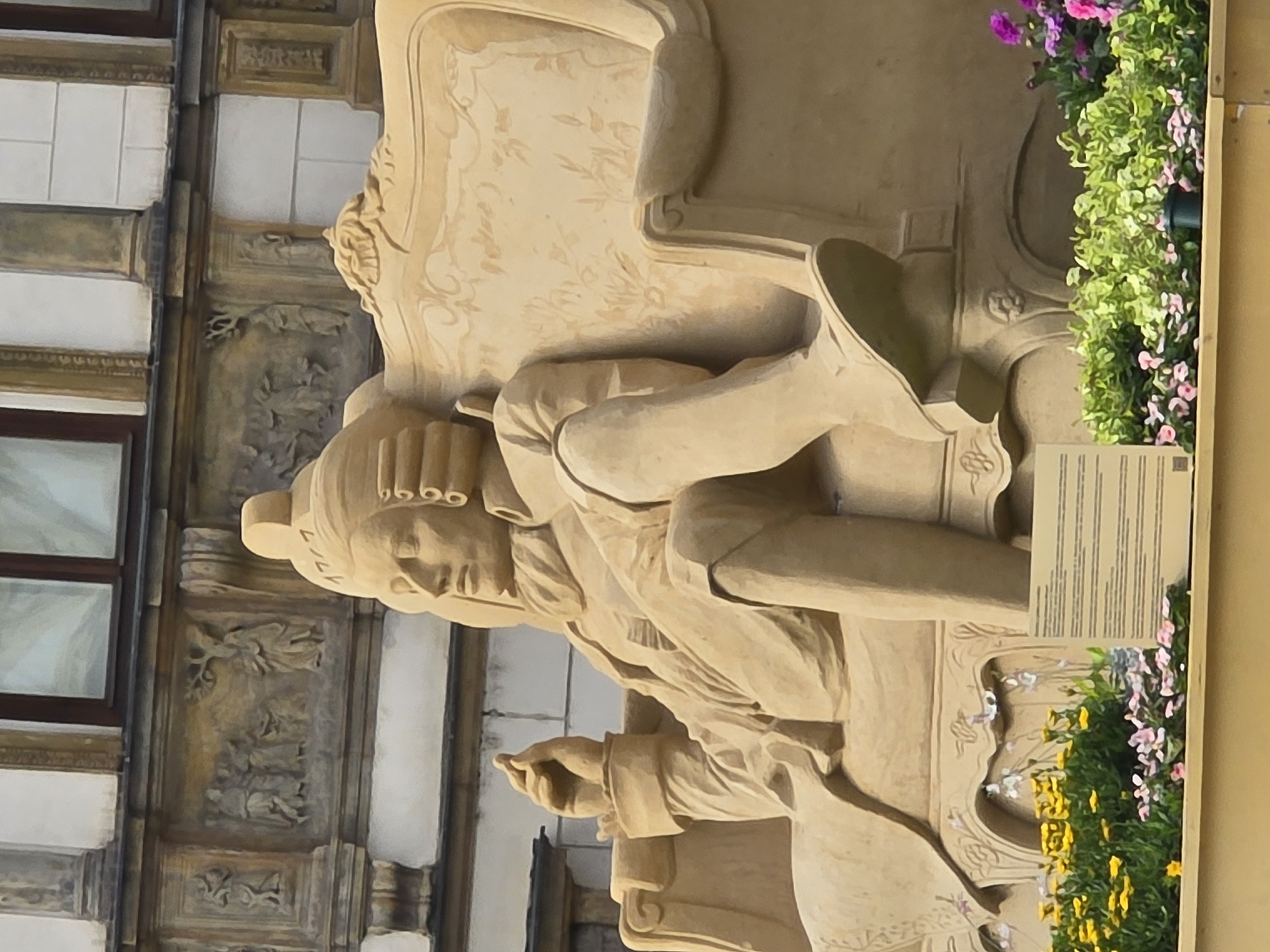 A large, elaborate sand sculpture depicts a seated man in historical attire, likely a nobleman or historical figure. He is positioned on an ornate armchair, with one leg casually crossed over the other. The figure is wearing a powdered wig, a ruffled cravat, and a long coat. His right hand is raised, and he appears to be holding something between his fingers, possibly a snuff box or a coin. The sand sculpture is placed in front of a building with classical architectural elements, including carved reliefs and columns. In the foreground, there is a display of colorful flowers, with yellow and pink blooms prominently featured. A small information plaque is visible near the flowers. The overall scene suggests a public art installation or historical reenactment, likely in Olomouc, Czechia, given the context of such sand sculptures often being featured in festivals or public spaces. The lighting indicates it is daytime, possibly sunny, as there are subtle shadows on the sculpture and surrounding architecture.