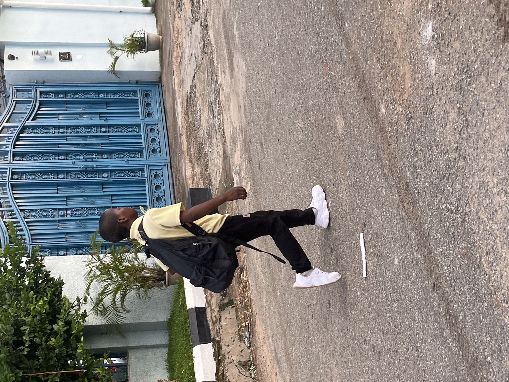 A young boy, appearing to be of African descent, walks right to left across a paved street. He is wearing a yellow short-sleeved shirt, black pants, and white sneakers. A black backpack is slung over his shoulders. He is walking alongside a curb with black and white markings, bordered by a patch of grass. Behind the curb, there are plants and a large, ornate blue gate. The gate is tall and appears to be made of metal, with intricate decorative elements. The wall behind the gate is light blue and extends to the right. To the right of the gate, a small potted plant sits on the ground. The street itself is asphalt, with some patches of wear and tear, and a white piece of paper or plastic is visible on the ground in the lower portion of the image. The scene suggests an outdoor urban environment, potentially in Abuja, Nigeria, given the style of the gate and the architecture.