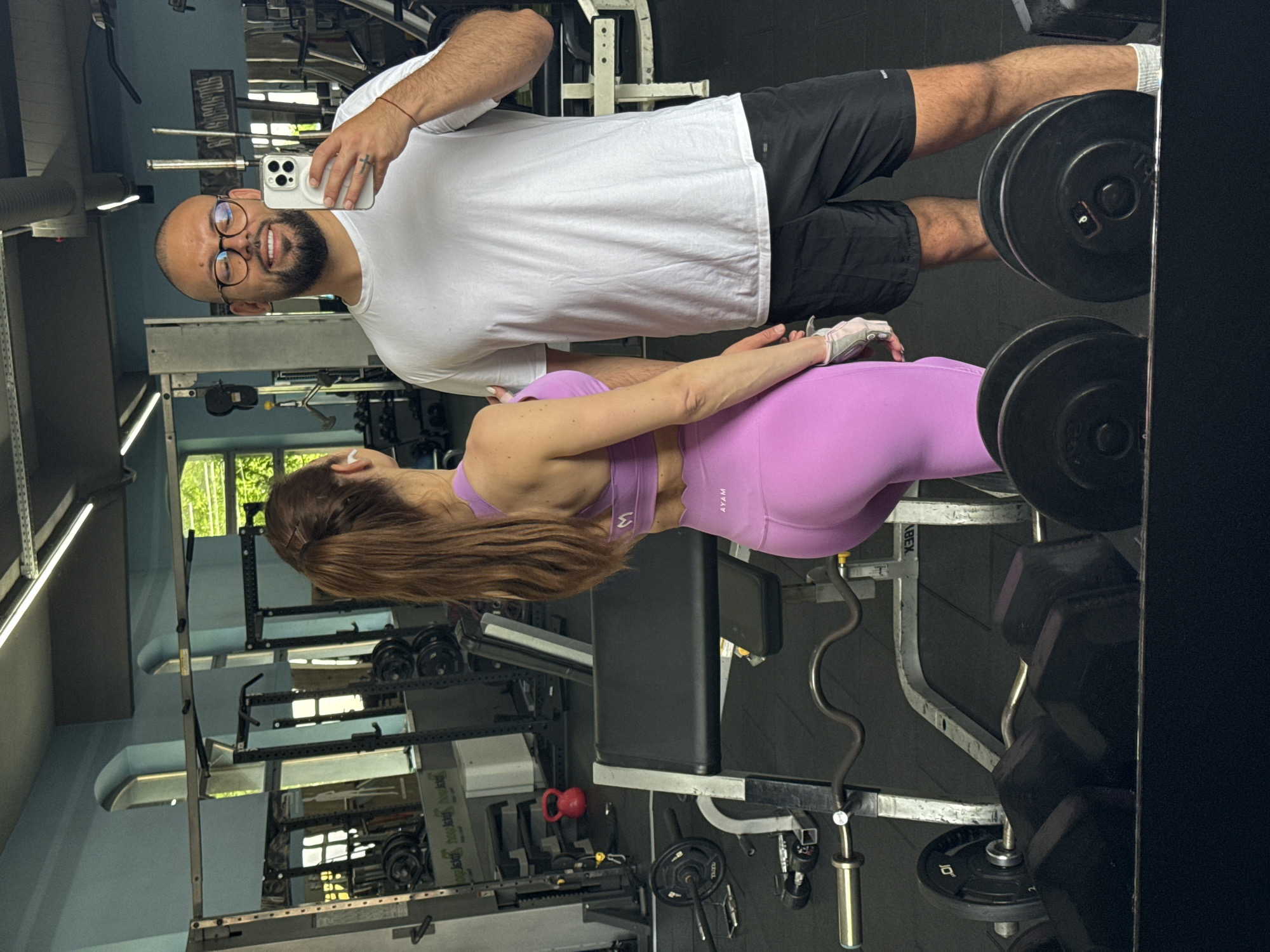 This mirror selfie captures a man and a woman inside a gym, likely in Plovdiv, Bulgaria, during what appears to be daytime. The man, positioned in the foreground and taking the photo with a white smartphone, stands to the right of the woman. He has a bald head, a dark beard, and is wearing glasses, a white t-shirt, and black shorts. Notable details include a ring on his left ring finger, a small cross tattoo on his right hand, and a red string bracelet on his left wrist. He is smiling broadly, holding the phone up to capture both himself and the woman.
The woman is standing facing away from the camera, looking towards the man, with her body angled slightly towards the right side of the frame. She has long brown hair tied back in a ponytail and is dressed in a light purple sports bra with a scalloped edge and matching purple leggings, which have the text "AYAM" visible on the hip. She is also wearing white fingerless gym gloves.
The setting is an indoor gym with various exercise equipment visible in the reflections. To the right of the man, a rack of black dumbbells is partially visible, with a weight plate showing the number "6". In the background, other gym equipment includes a squat rack (visible with mirrored text "SQUAT CON..." and "SQUAT RACK"), benches, and a red kettlebell. Natural light streams in from windows, reflecting off the mirror. The overall mood suggests a relaxed moment during or after a workout.