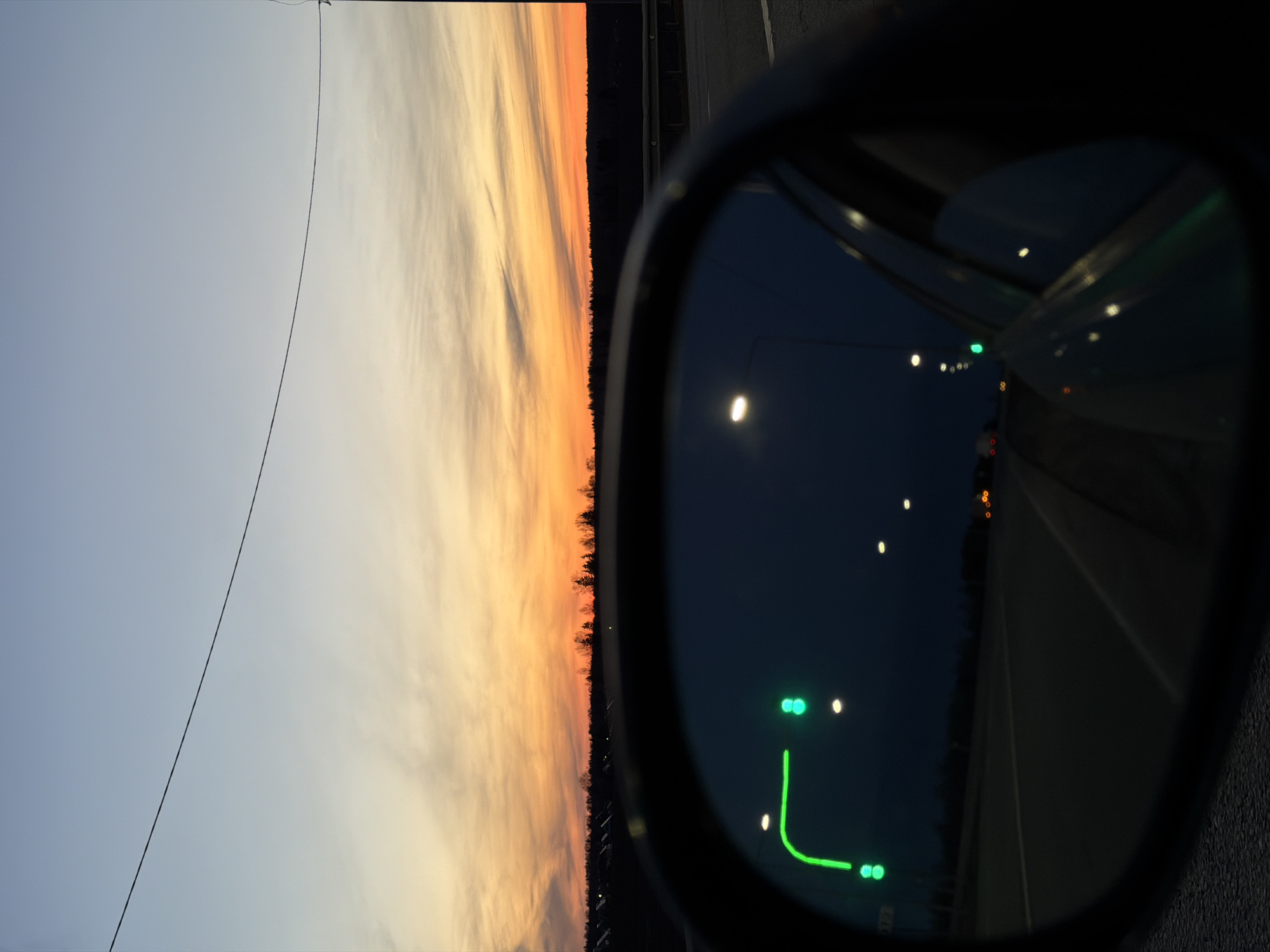 The image presents a dual scene: a sunset vista contrasted with the reflection in a car's side mirror. The sunset displays a gradient of colors, from light blues/grays to oranges/reds, likely indicating the end of the day. A tree line obscures the horizon beneath the sunset. A power line traverses the sky.
The car's side mirror reflects a dark road with streetlights and possibly traffic signals, including a set of two green lights and an artificial turn signal. The scene within the mirror has a distinctly nighttime or very late evening ambiance.
There is nothing in the image to confirm this location in the city of Kuregovo, Russia.