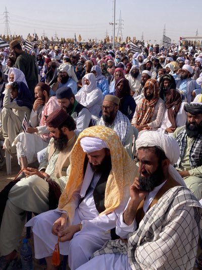 This media file captures a large outdoor gathering of men in Pishin, Pakistan, under a clear, bright sky. The scene is set in a dry, open area with sandy or dusty ground. The crowd consists predominantly of adult men, many with beards, dressed in traditional South Asian attire, including shalwar kameez, various forms of head coverings such as white turbans, patterned scarves, and different styles of caps.
The men are densely packed, with some seated on white plastic chairs in the foreground and middle ground, while others appear to be standing or seated directly on the ground further back. In the distance, a vast number of people stretch across the landscape. Several individuals are seen holding small black and white striped flags, which are also visible prominently throughout the crowd in the background, suggesting that this is an organized event, possibly a political rally or a religious congregation associated with a particular group or party.
Notable background elements include large electricity transmission towers (pylons) under a clear sky, indicating sunny weather conditions. The overall atmosphere appears calm and attentive, with most individuals looking forward, seemingly listening to a speaker or observing a focal point outside the frame. Some men are seen holding prayer beads (tasbih), and a plastic water bottle is visible on the ground next to one of the seated men. No discernible text is visible on flags or other objects.