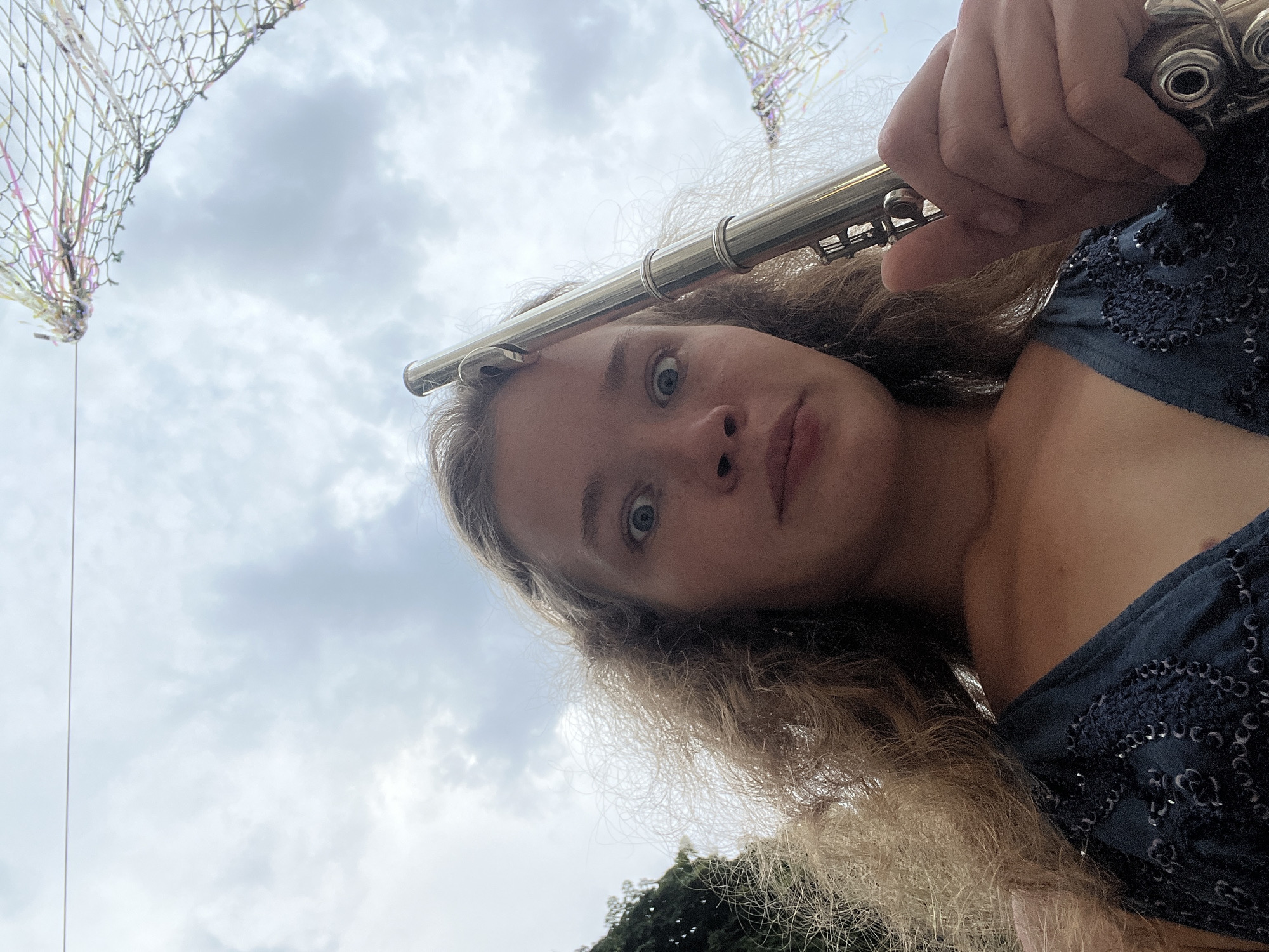 In Budva, Montenegro, a young woman with long, wavy blonde hair, dressed in a dark blue, sequined top, looks directly at the camera with wide, surprised eyes. She is holding a silver flute vertically against her forehead with her right hand. The background consists of a cloudy sky and some decorative netting with colorful ribbons hanging from it. A dark green tree is visible on the left side of the frame.