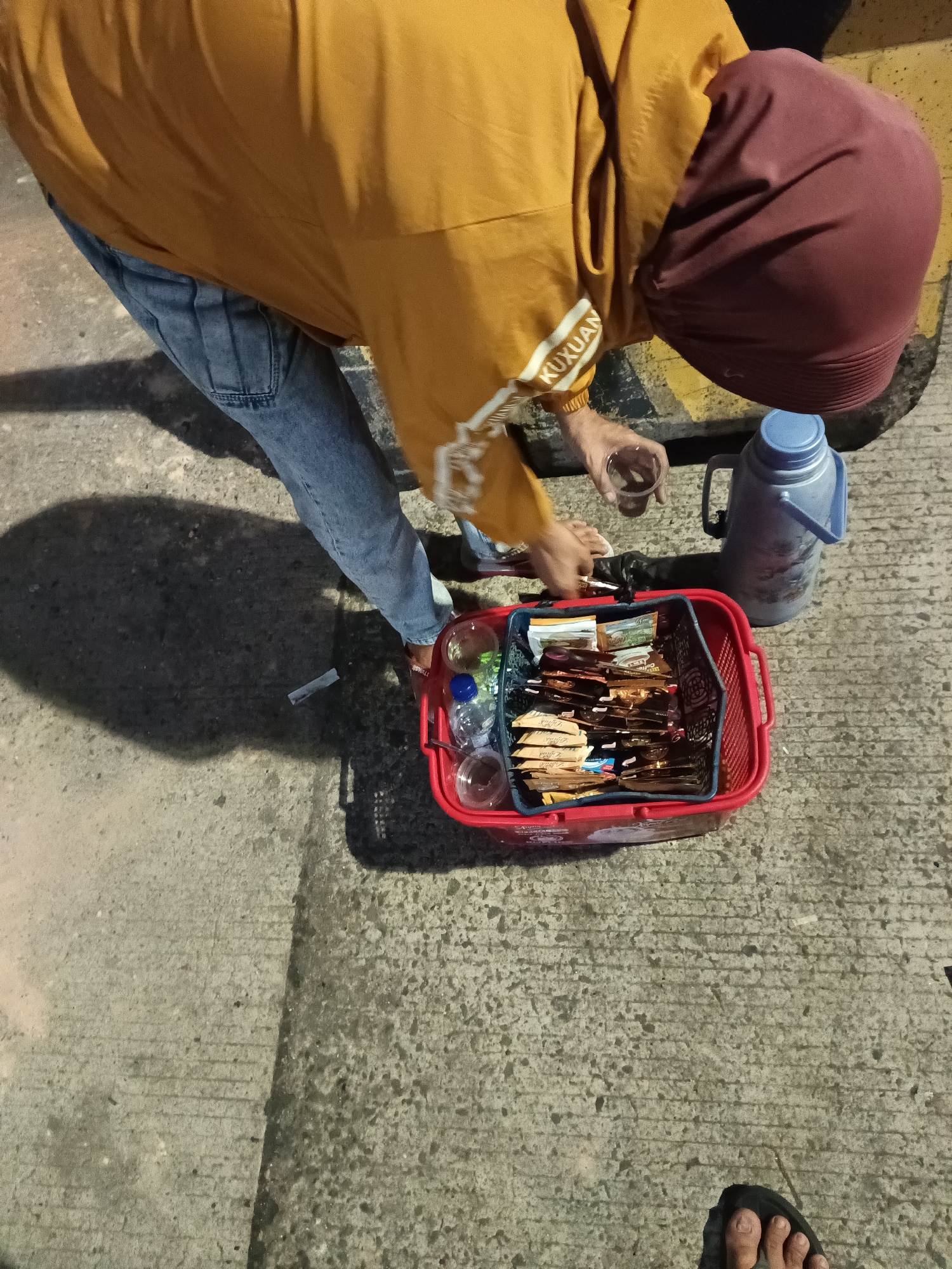 A person wearing a yellow jacket, blue jeans, and a burgundy headscarf is bent over a red basket filled with small packets of food items, likely snacks or instant beverages. The basket also contains a few plastic bottles and cups. To the right of the basket is a tall, silver thermos. The person is holding a clear cup with some liquid in it. The scene appears to be on a concrete surface, possibly a sidewalk or road, with visible lines and textures in the concrete. The lighting suggests it might be evening or night, casting shadows from the person and the basket. The image has been identified as being taken in Bakauheni, Indonesia.