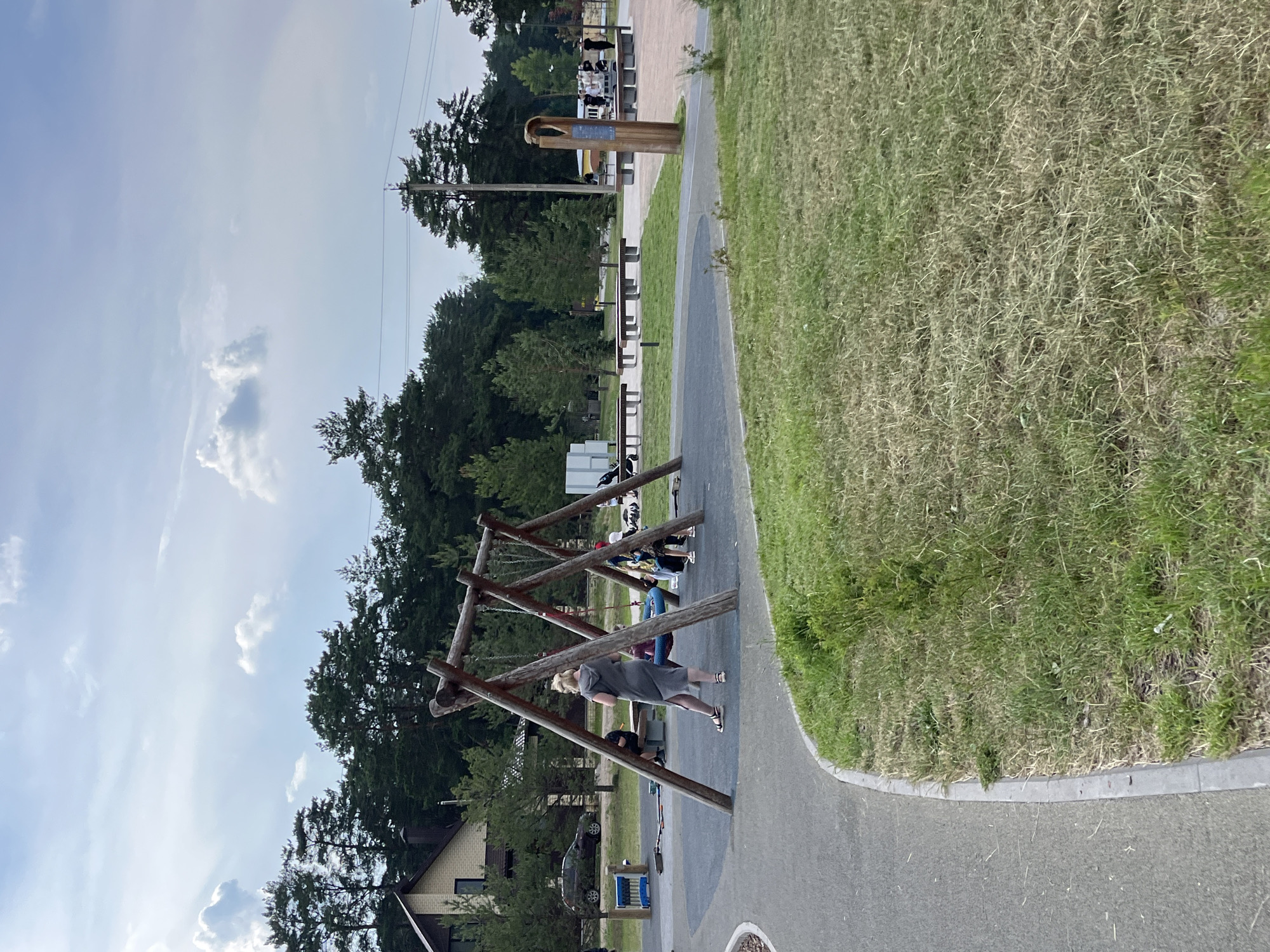 This image depicts an outdoor children's playground area in Zelenodol'sk, Russia. A large wooden swing set with two swings is a central feature, with several individuals visible nearby. One person, wearing a grey hooded dress and sandals, stands near the swing set, facing away from the camera. Two children are seated on one of the swings. In the background, a row of benches is visible, some occupied by people. To the left, a building with a brown roof and light-colored walls is situated near a dark-colored car. The playground is surrounded by a paved pathway and grassy areas with scattered trees, predominantly conifers, and a dense forest line further in the distance. The sky is partly cloudy.