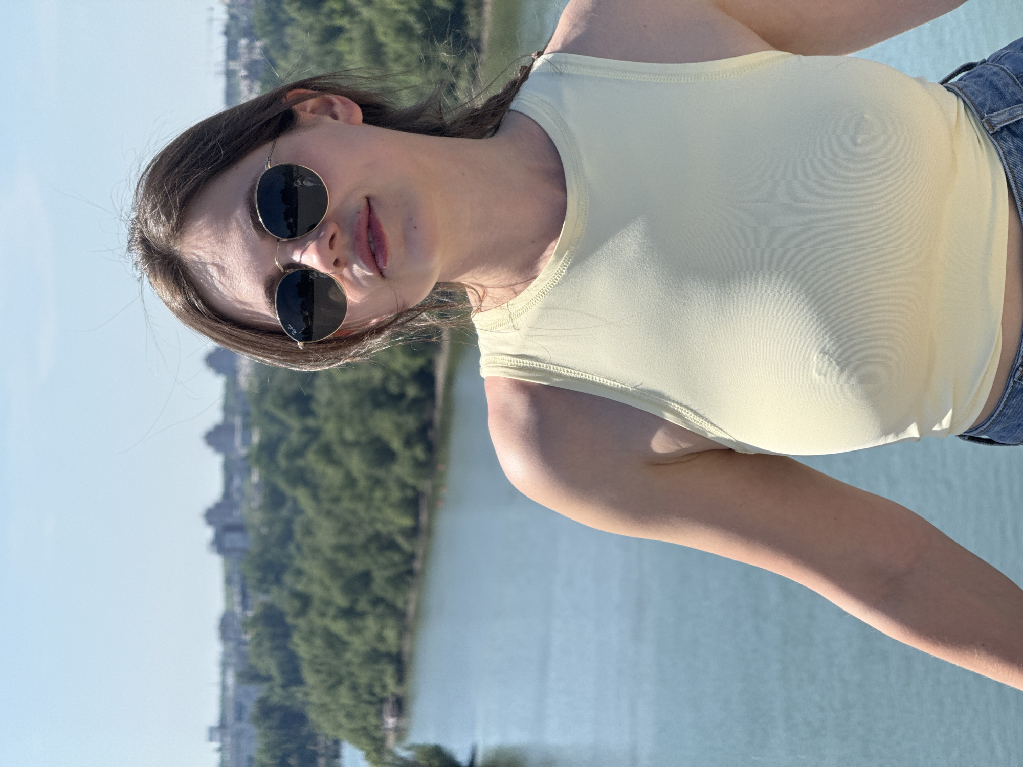 A woman wearing sunglasses and a yellow tank top stands on a bridge overlooking the Sava River in Belgrade, Serbia. Trees line the riverbank, and buildings can be seen in the distance. The sky is blue.